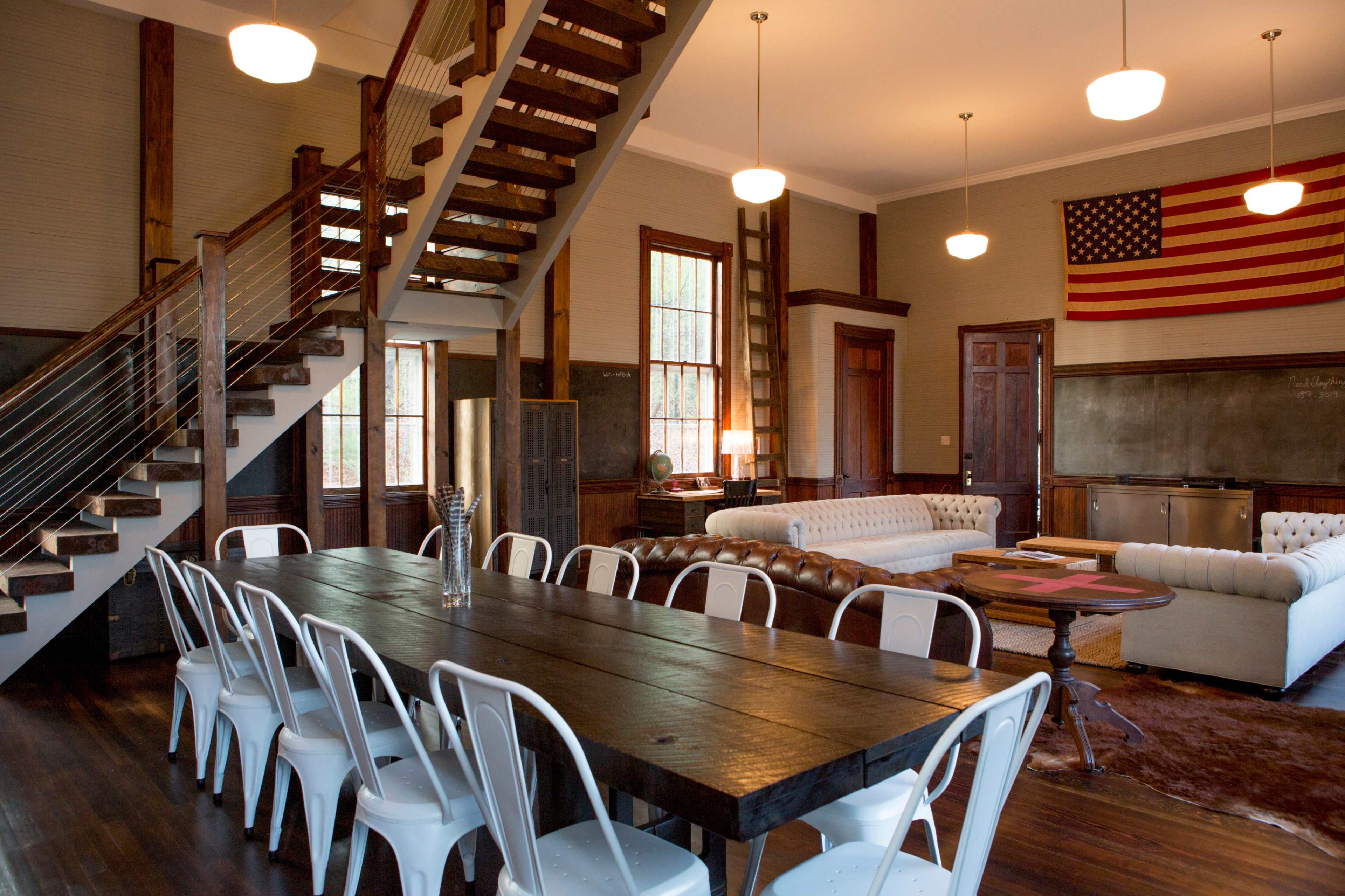 A spacious room features a long wooden dining table surrounded by metal chairs, with a staircase in the background and an American flag hanging on the wall.
