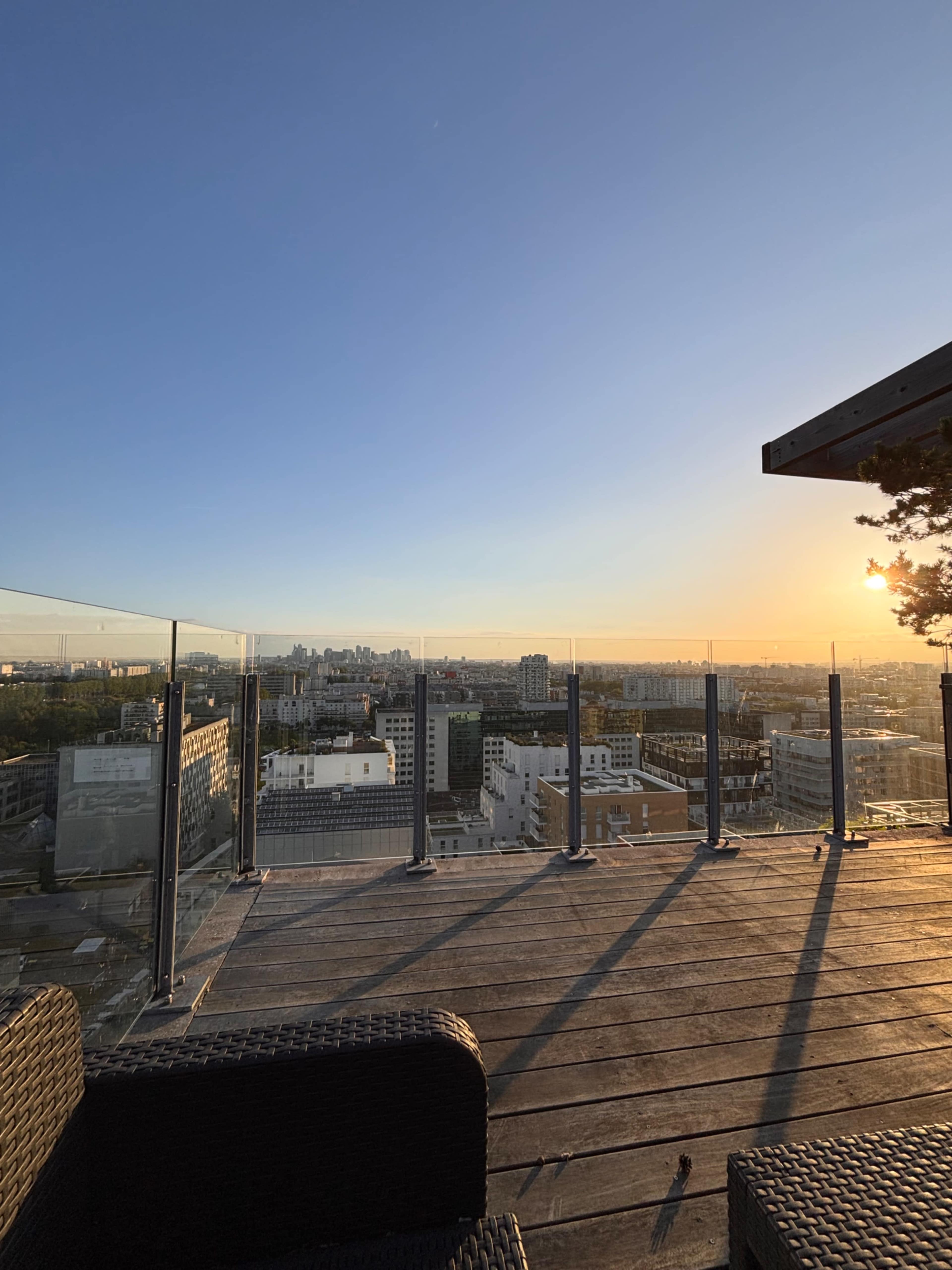 The image shows a rooftop terrace with glass railings, overlooking a city skyline at sunset.