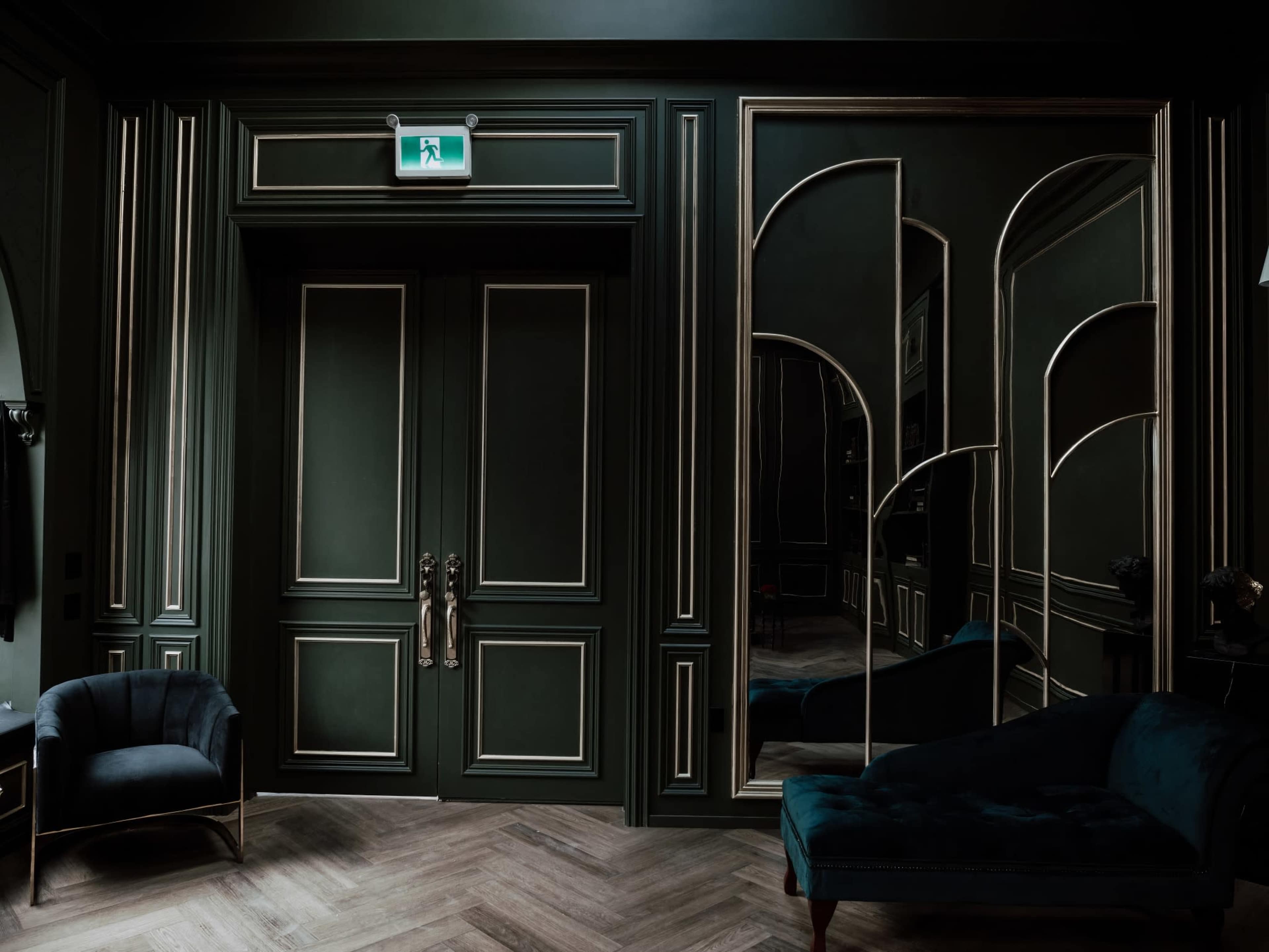 A dark green, elegantly designed room with a large mirror, double doors, and a velvet armchair.