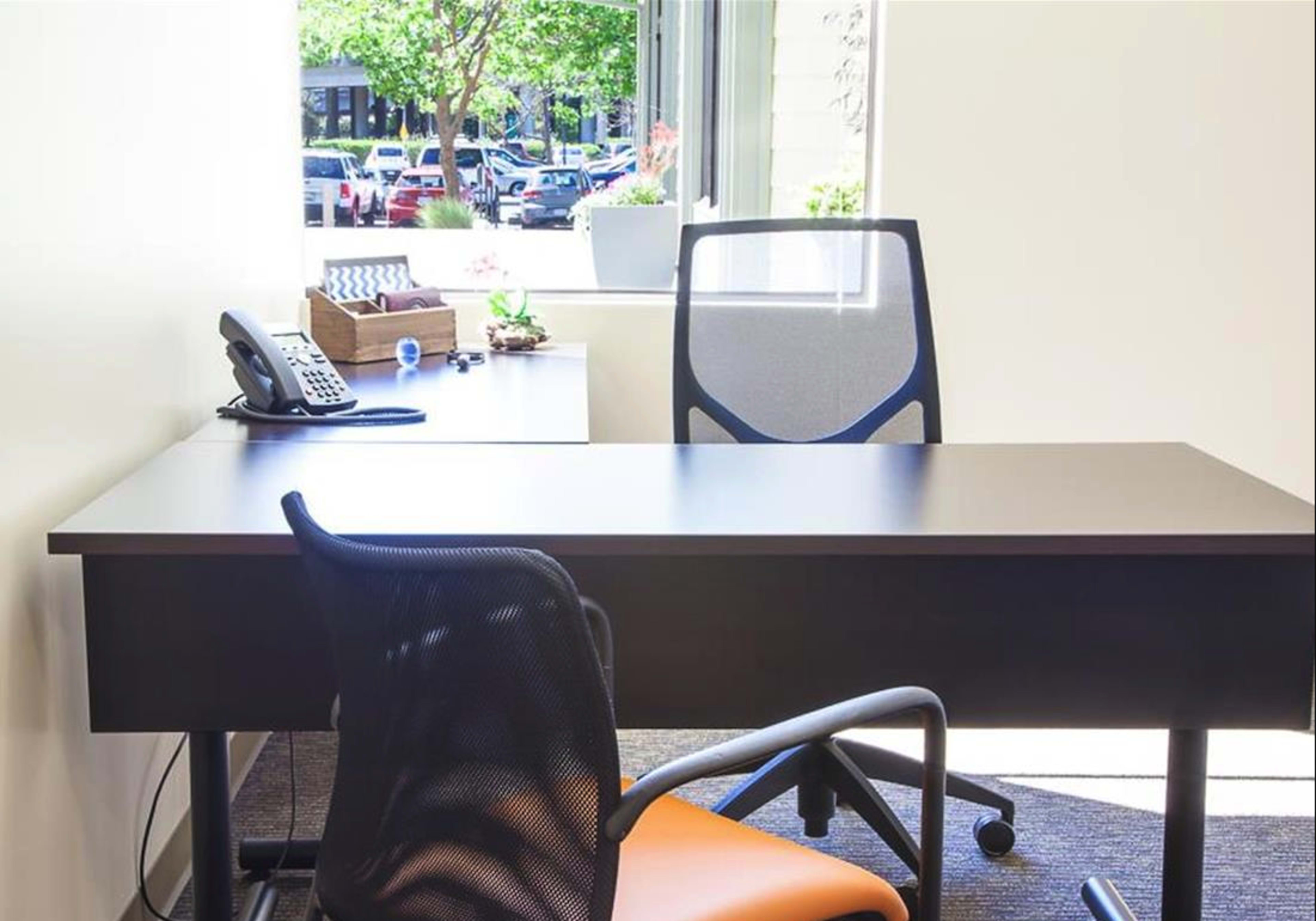 An office space with a desk, a chair, a phone, and a window overlooking a parking lot and greenery outside.