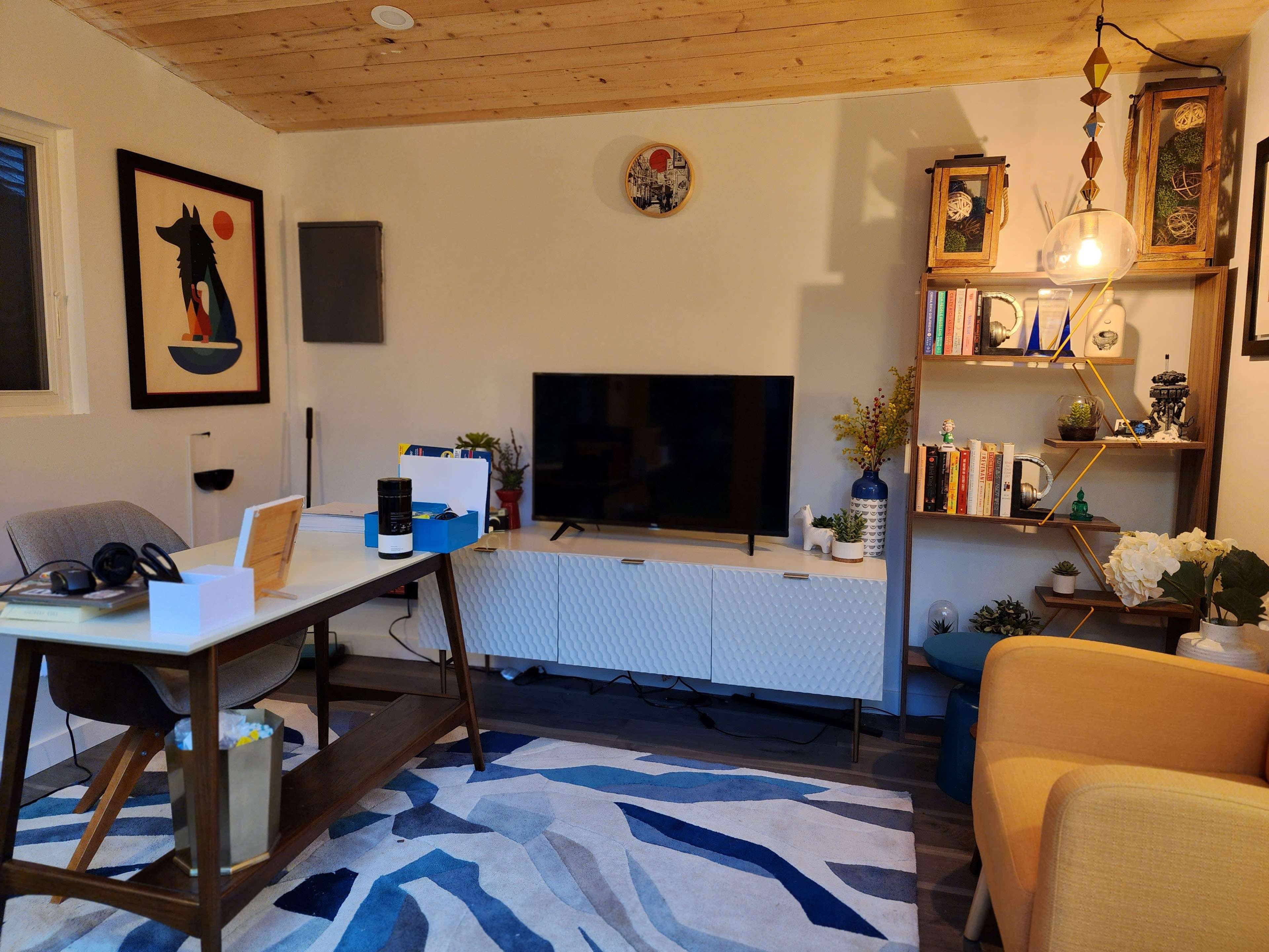 A contemporary living room with a television on a white media console, a wooden desk, an orange armchair, and various decorative items on shelves.