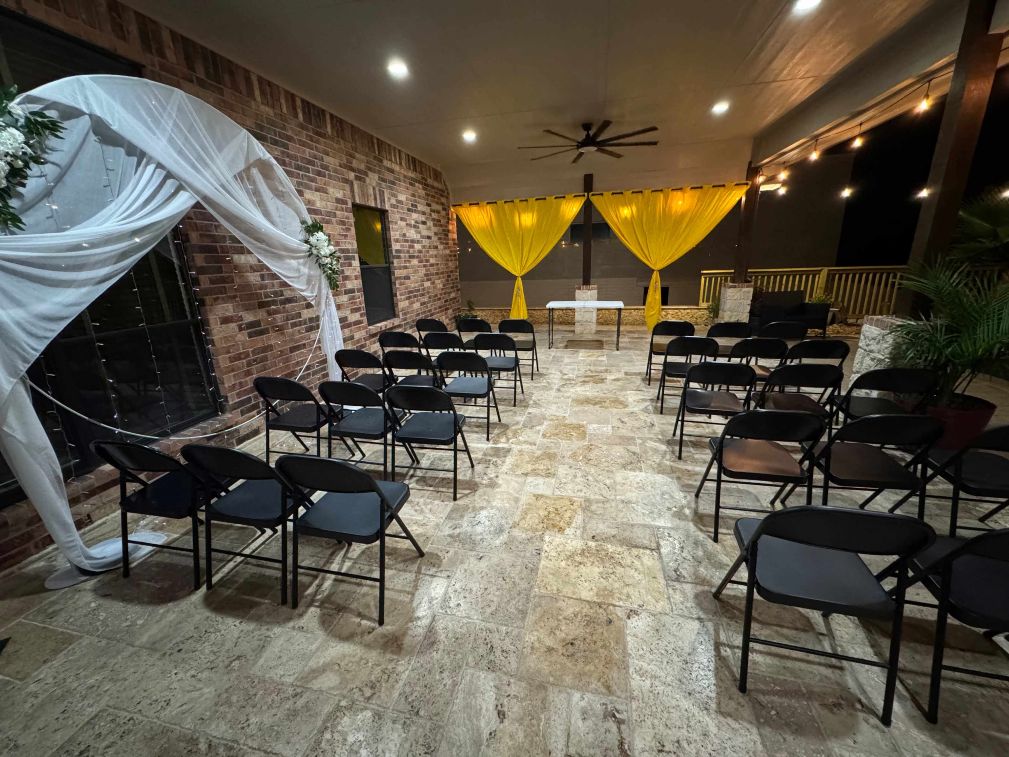 The image shows a dimly lit indoor venue with black chairs arranged in rows facing a decorated altar featuring white drapery and yellow curtains.