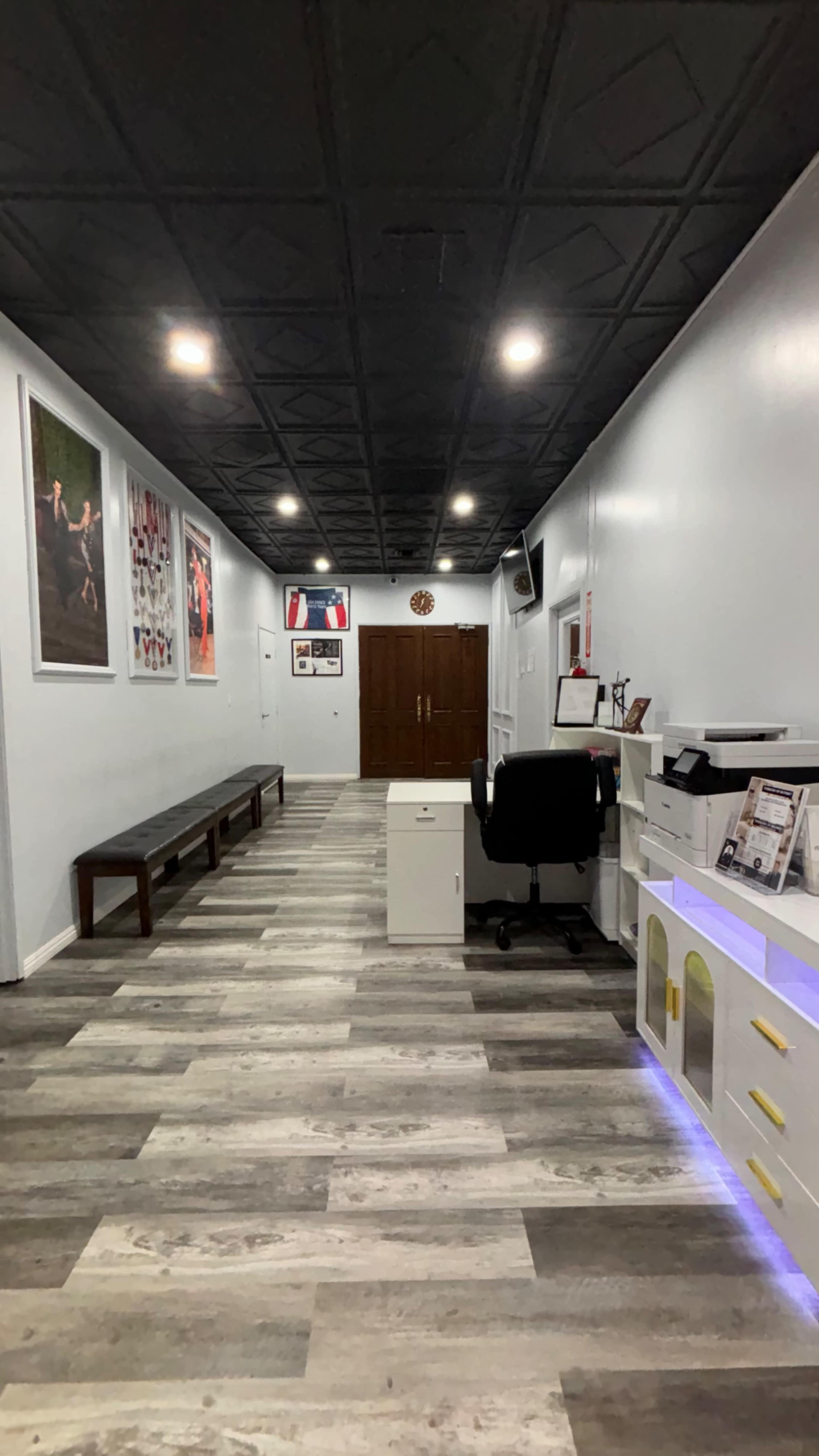The image shows a narrow hallway with a bench along one side, office furniture at the end, and framed photos on the walls.