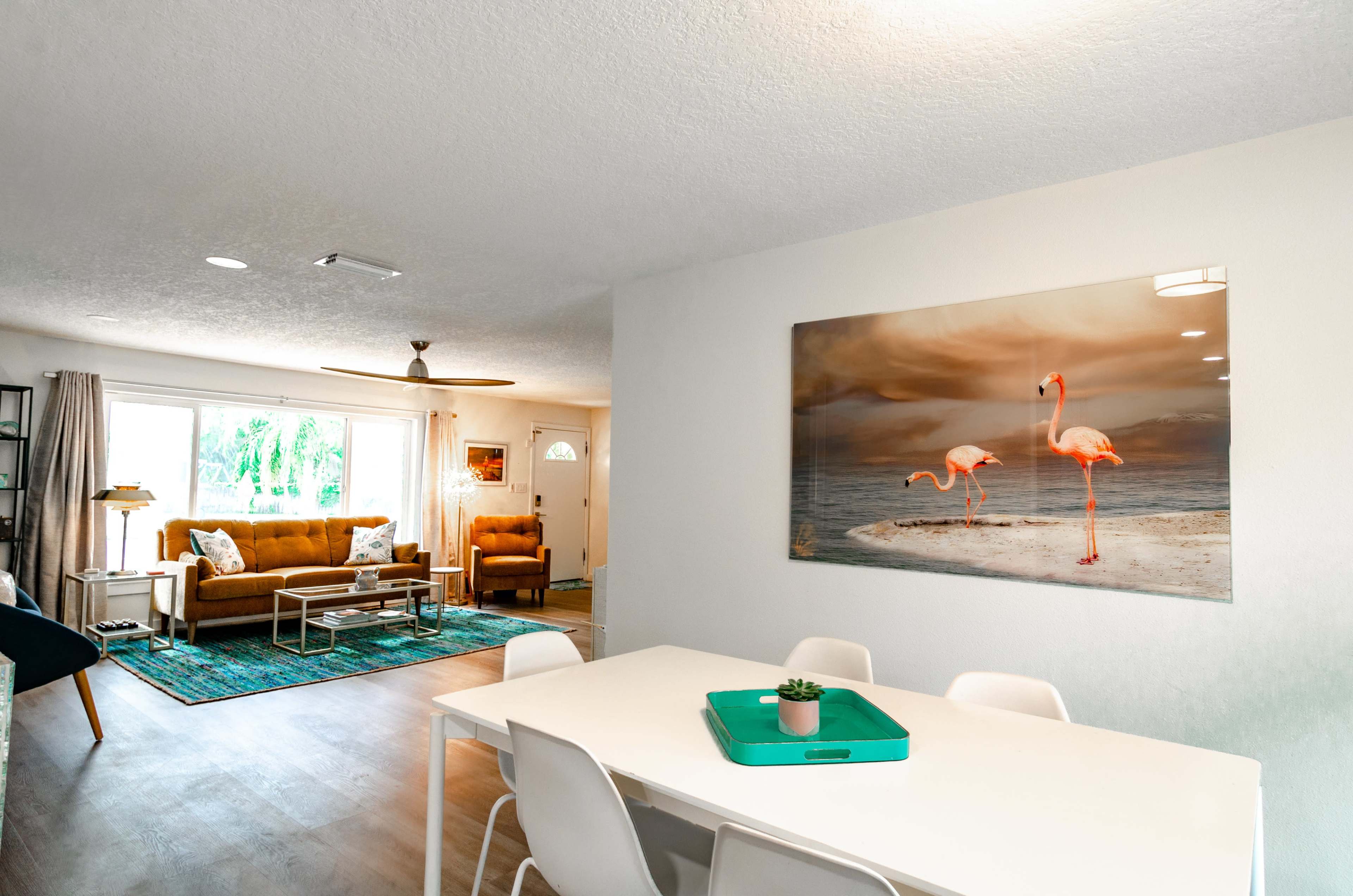 A spacious living area features a dining table with white chairs, a sofa, and a large artwork of flamingos on the wall.