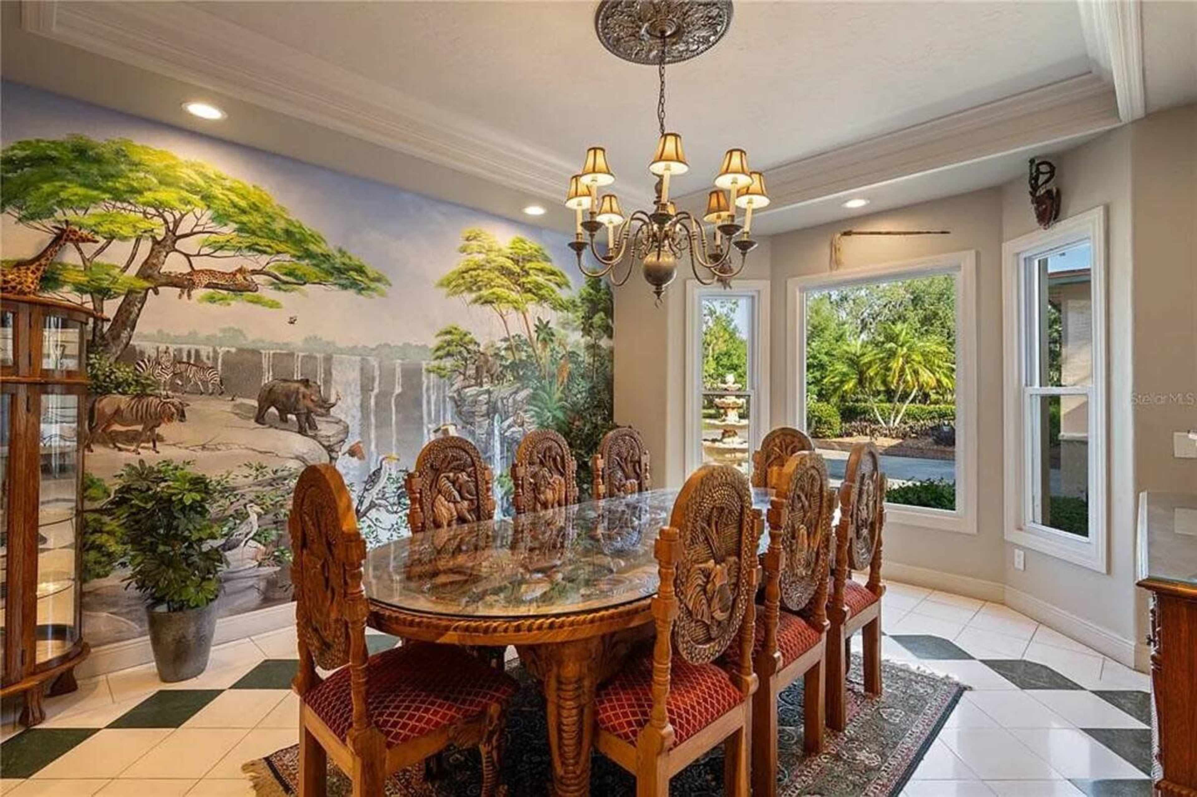 A dining room features a large wooden table surrounded by intricately carved chairs, with a mural of an African landscape on one wall and large windows showcasing greenery outside.