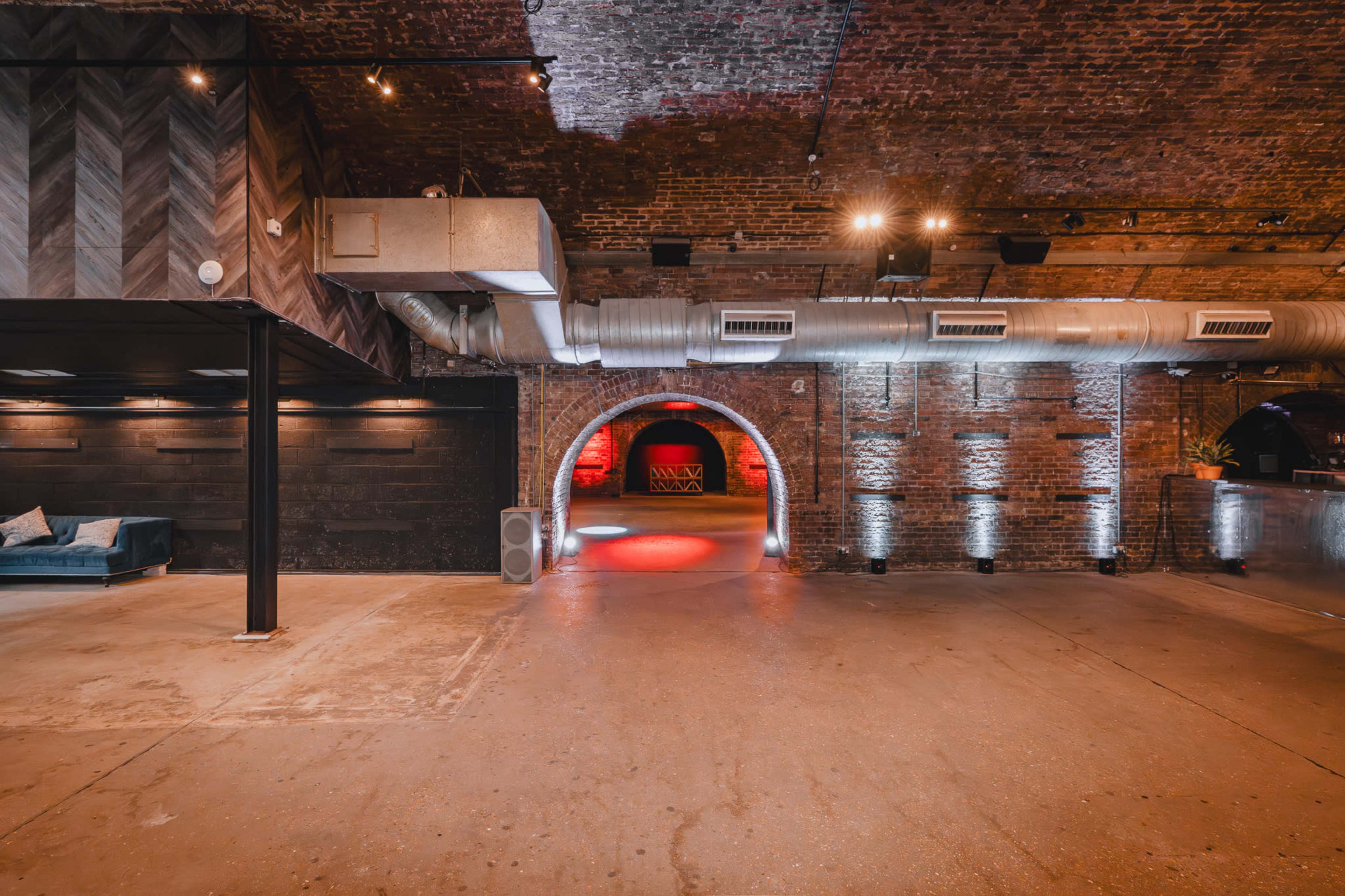 The image shows a spacious interior with exposed brick walls, a high ceiling, and industrial-style lighting, featuring a large archway illuminated with red light at the far end.