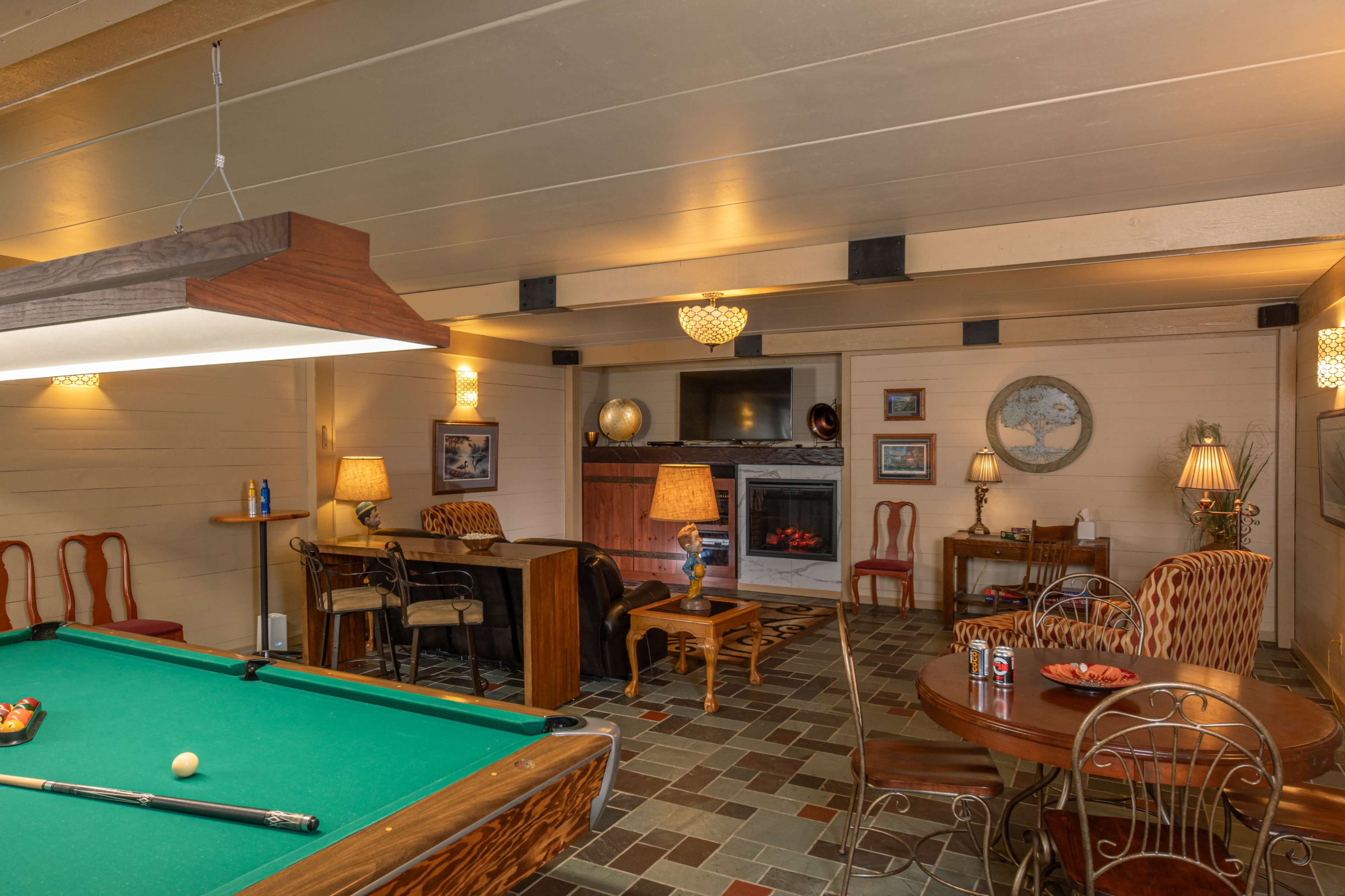 The image depicts a cozy game room featuring a pool table, a seating area with chairs and a table, and multiple lamps illuminating the space.