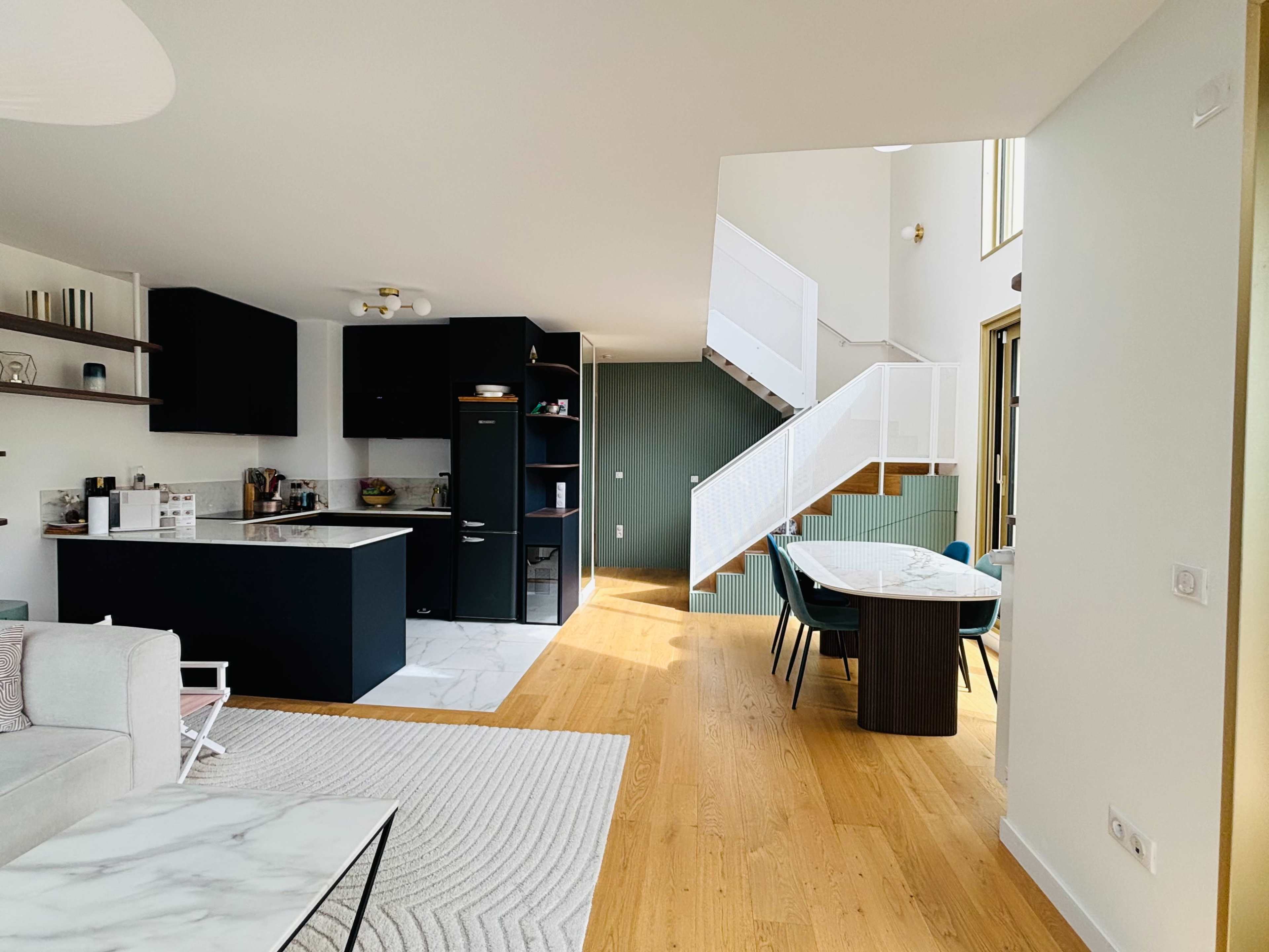 The image shows a modern, open-concept living space featuring a kitchen, dining area, and staircase, with wooden flooring and a mix of dark cabinetry and light decor.