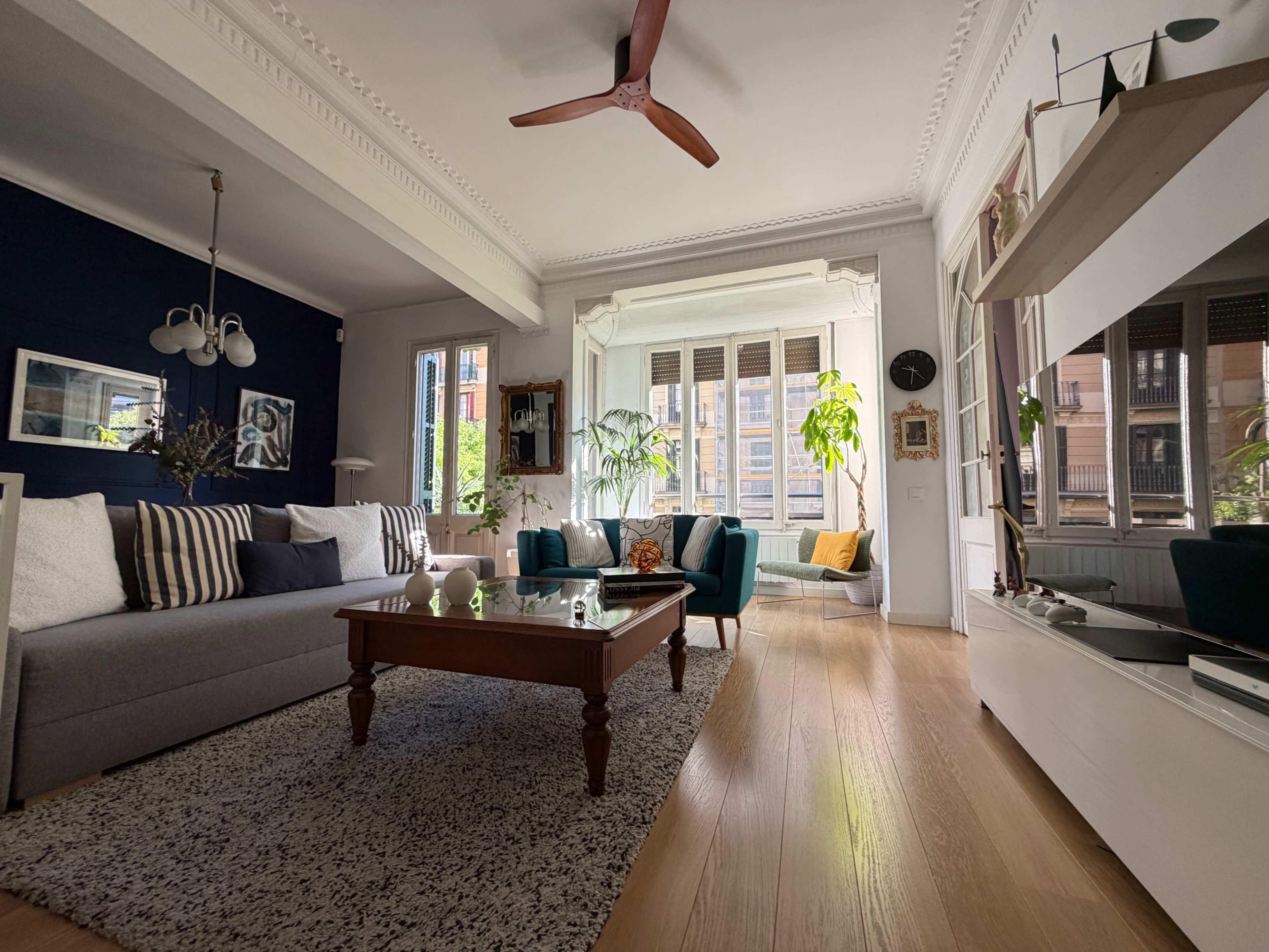 The room features a gray sofa, a wooden coffee table, and teal armchairs, with large windows allowing natural light to fill the space.