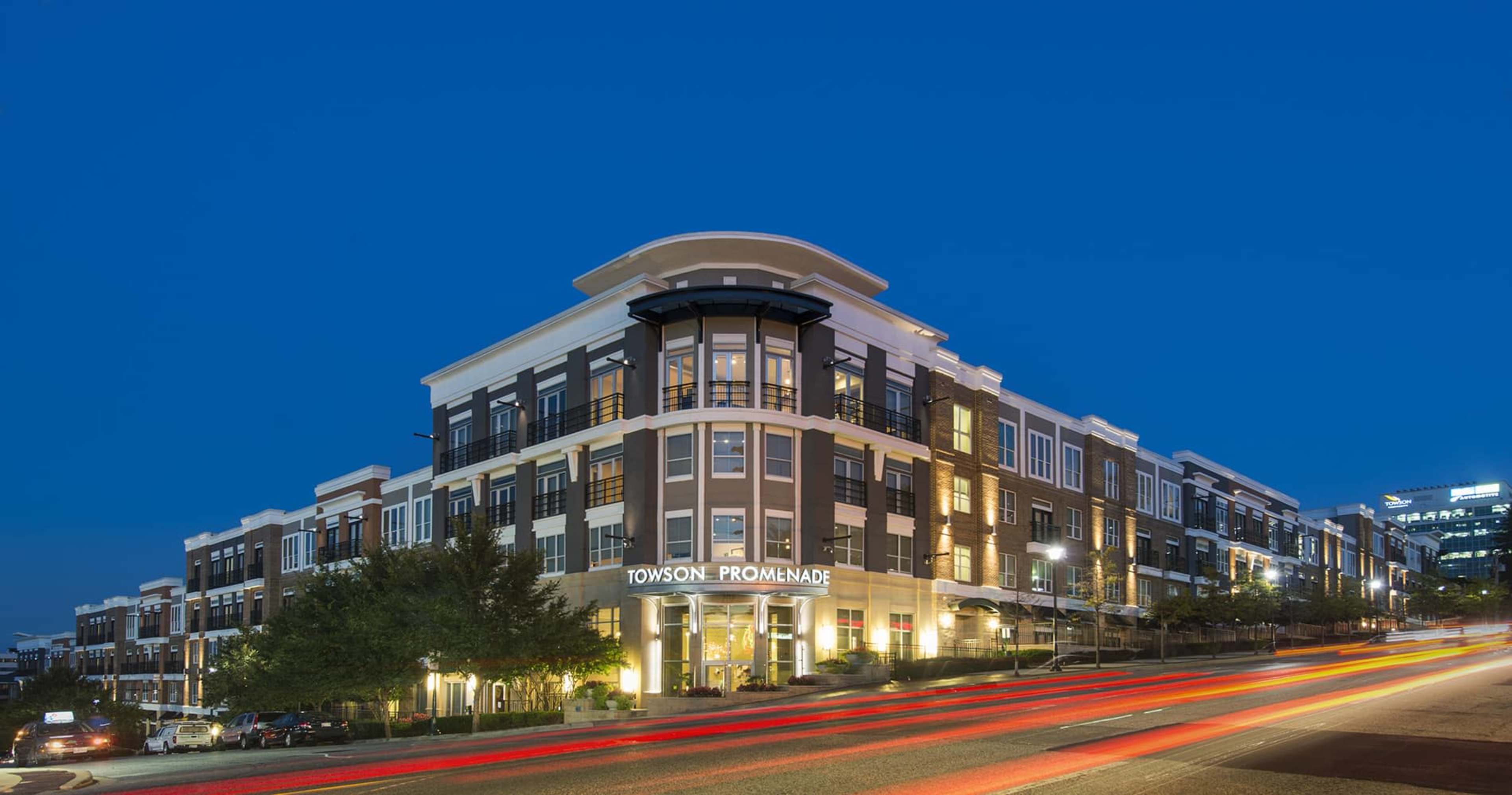 The Towson Promenade building illuminated at night, with streaks of car lights in the foreground.
