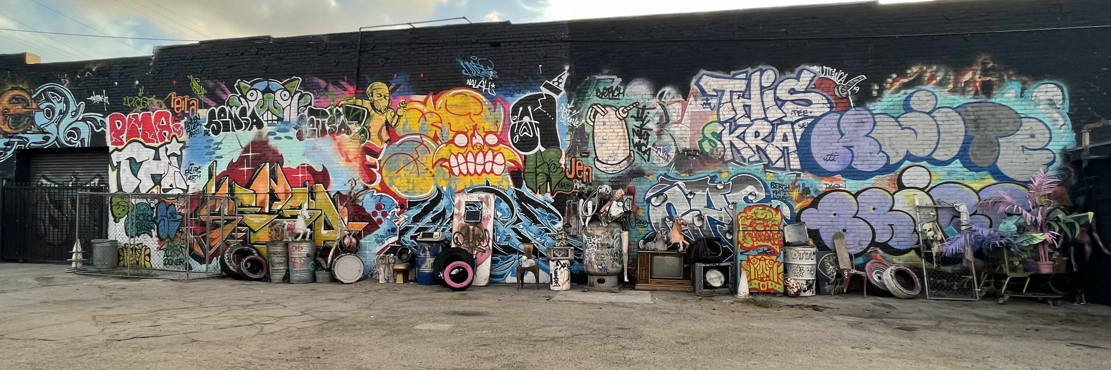 A long wall covered in colorful graffiti art is accompanied by various discarded objects, including tires and old appliances, arranged in front of it.