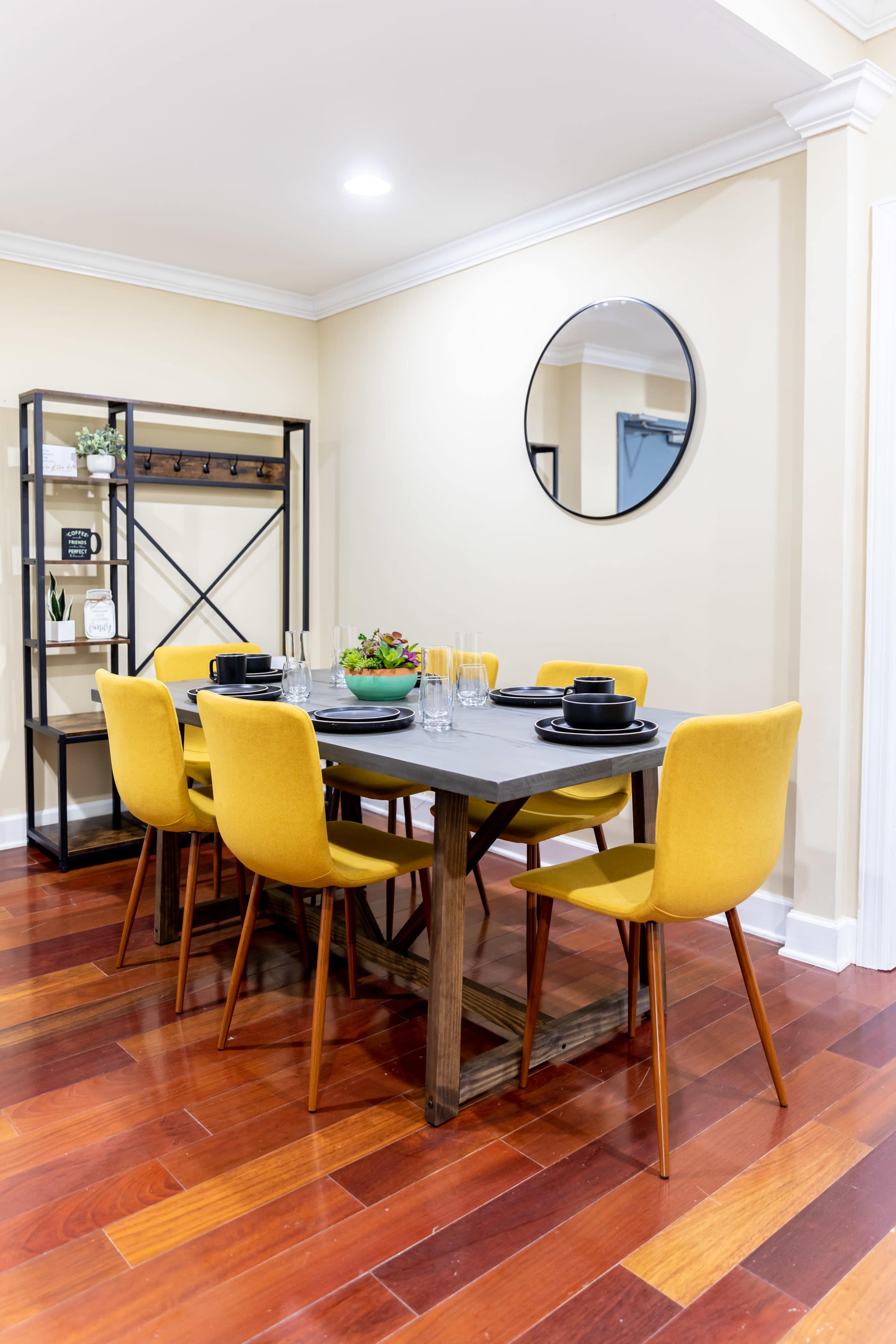 A dining area features a rectangular wooden table set for six with yellow chairs, a circular mirror on the wall, and a shelving unit in the background.
