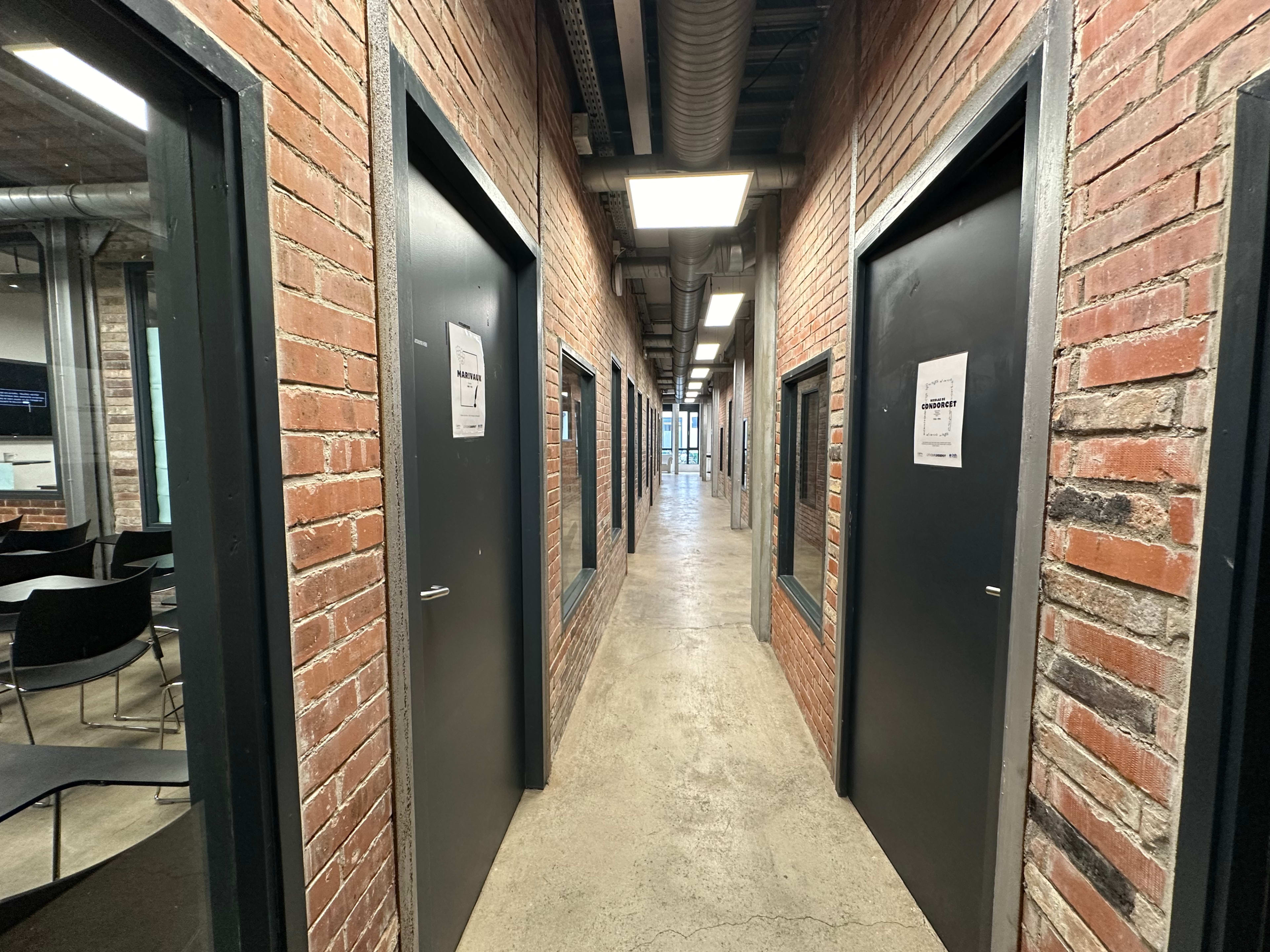 A narrow corridor lined with dark doors and exposed brick walls, leading to a well-lit area at the end.