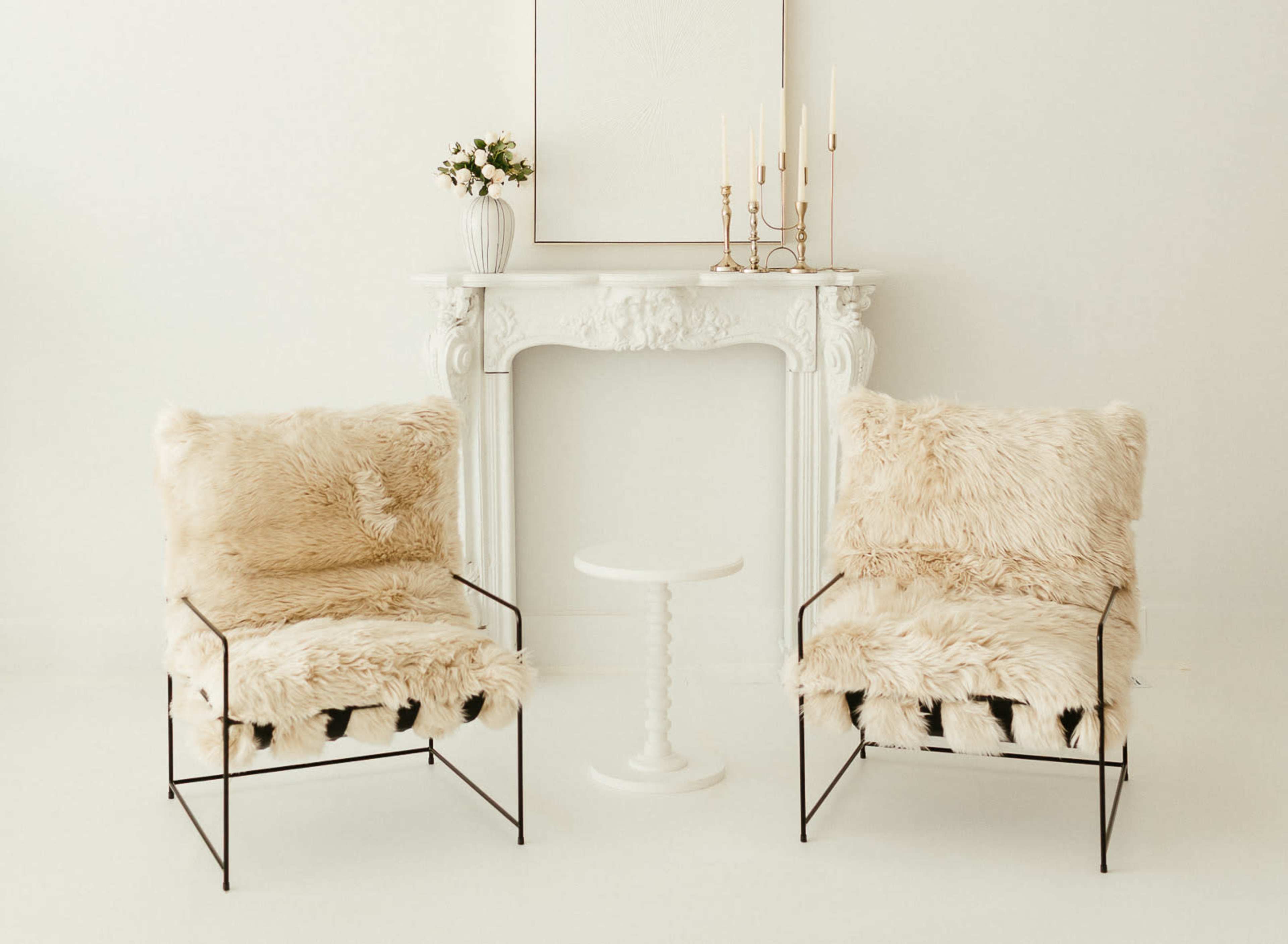 Two upholstered chairs with fur throws flank a small round table in front of a white mantle adorned with candles and a vase of flowers.