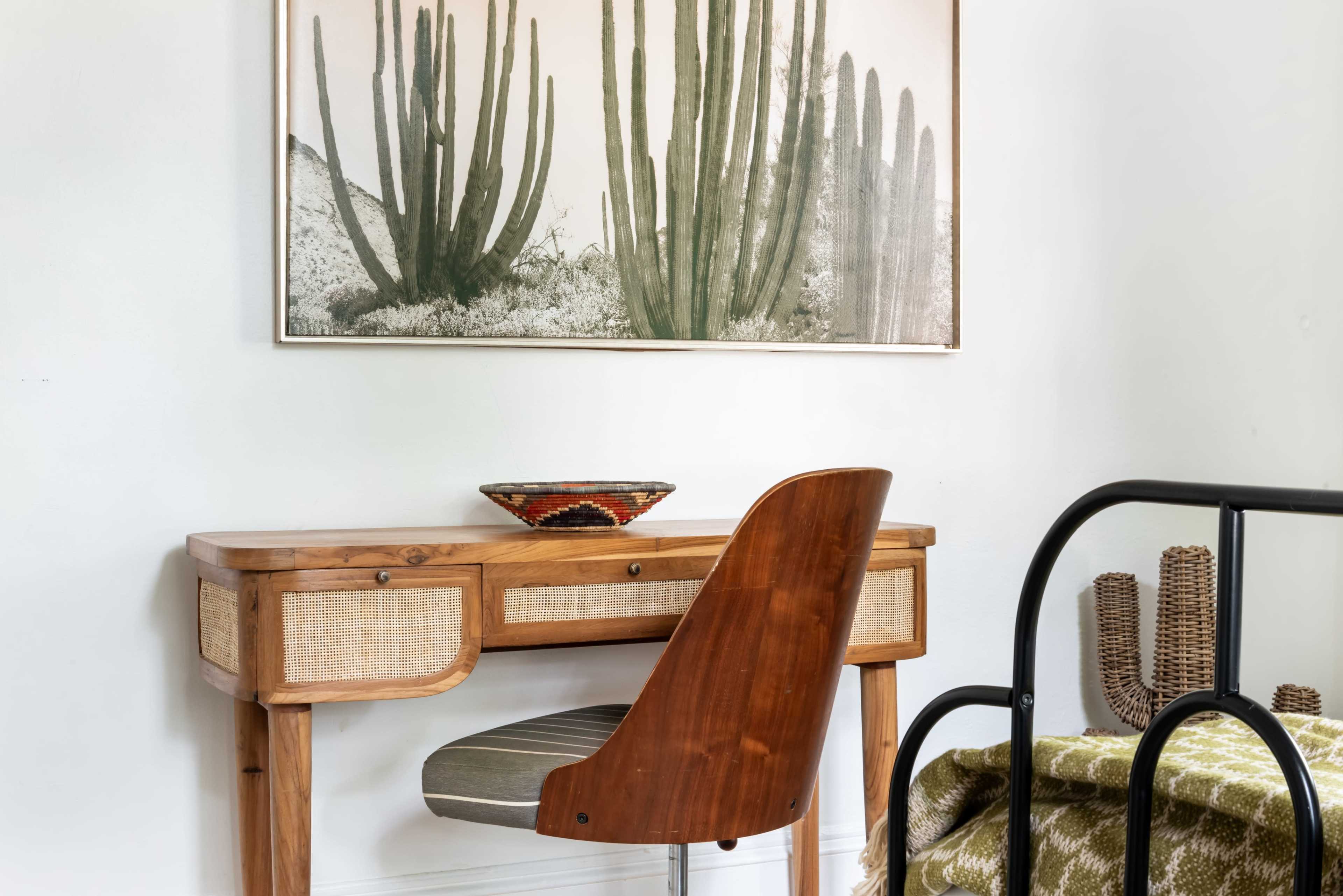A wooden desk with a chair is positioned next to a bed, complemented by a large cactus-themed artwork on the wall and a decorative bowl on the desk.