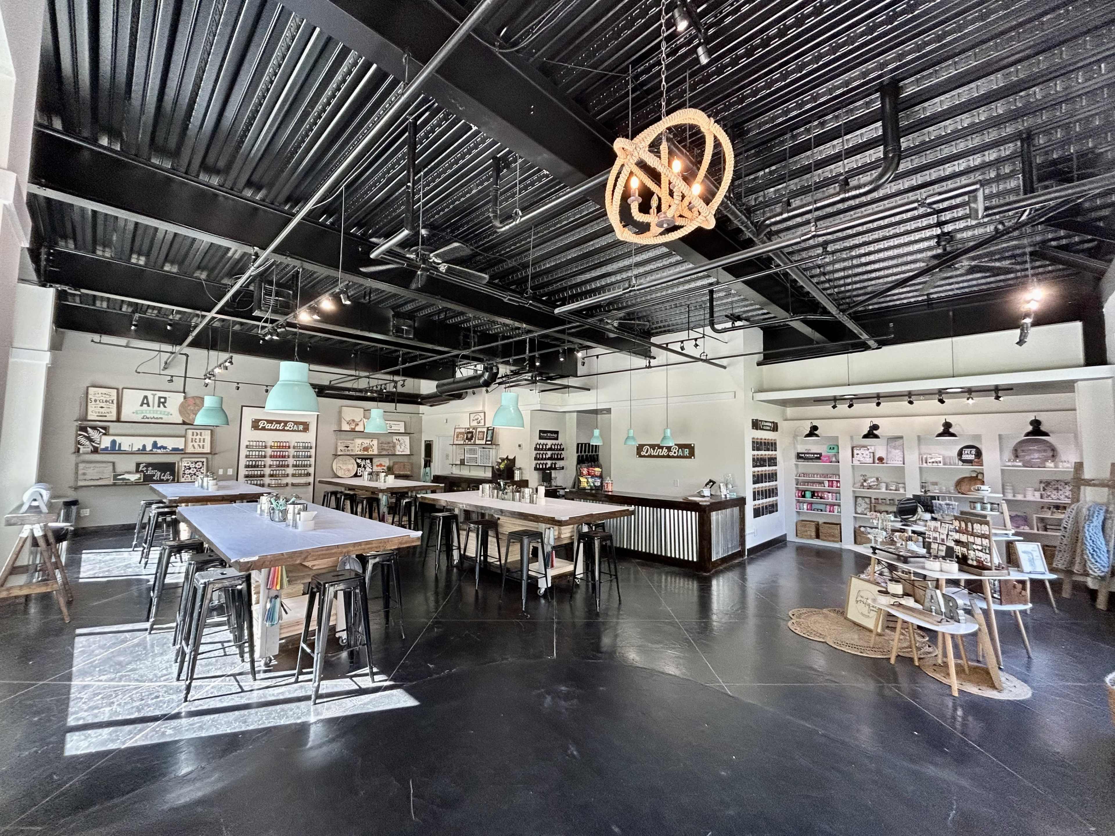 An open retail space with wooden tables, metal chairs, and shelves displaying various products, illuminated by pendant lights from a high ceiling.
