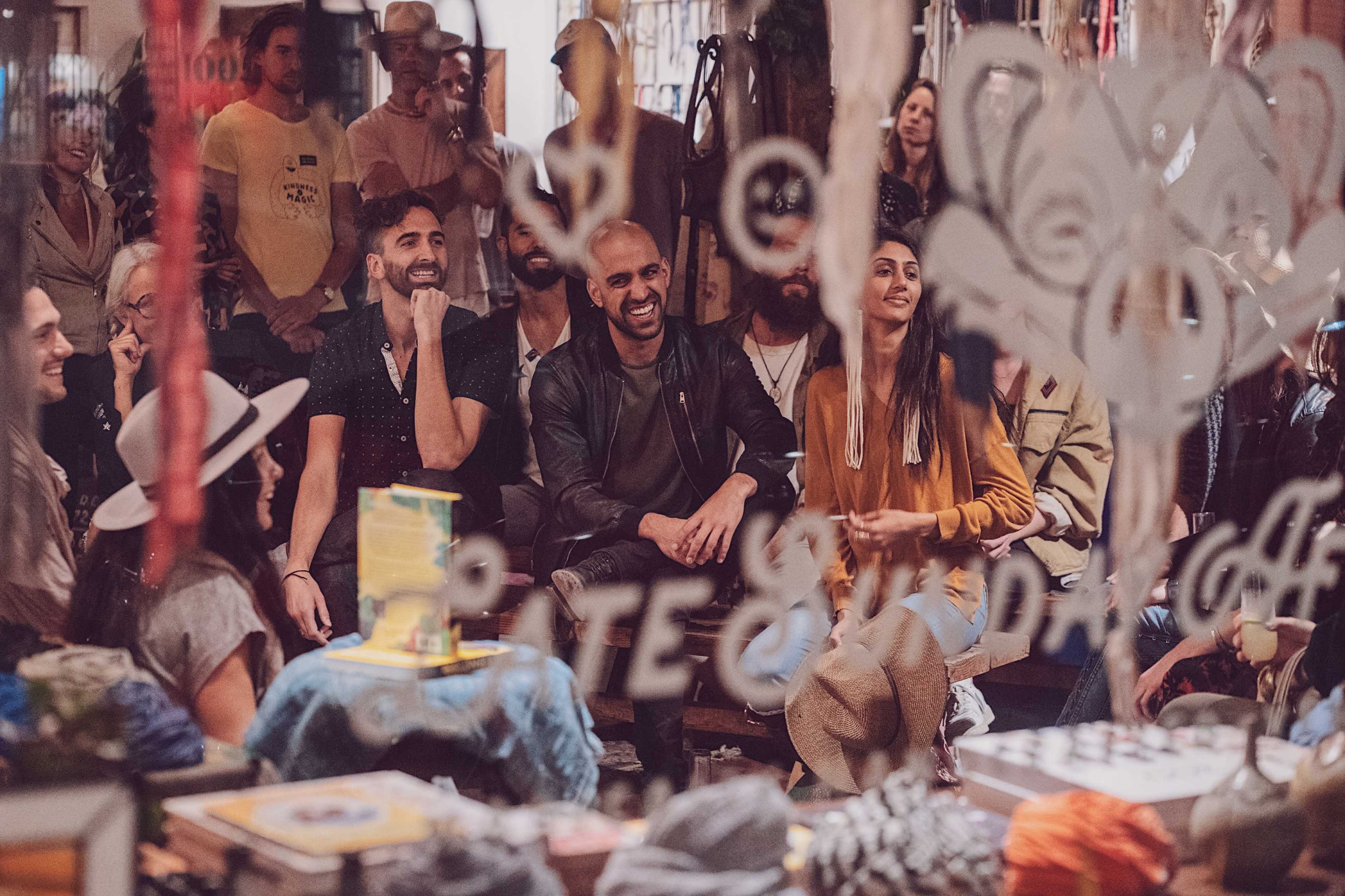 A diverse group of people is seated indoors, engaged in conversation and looking towards a focal point, with colorful items and decorations surrounding them.