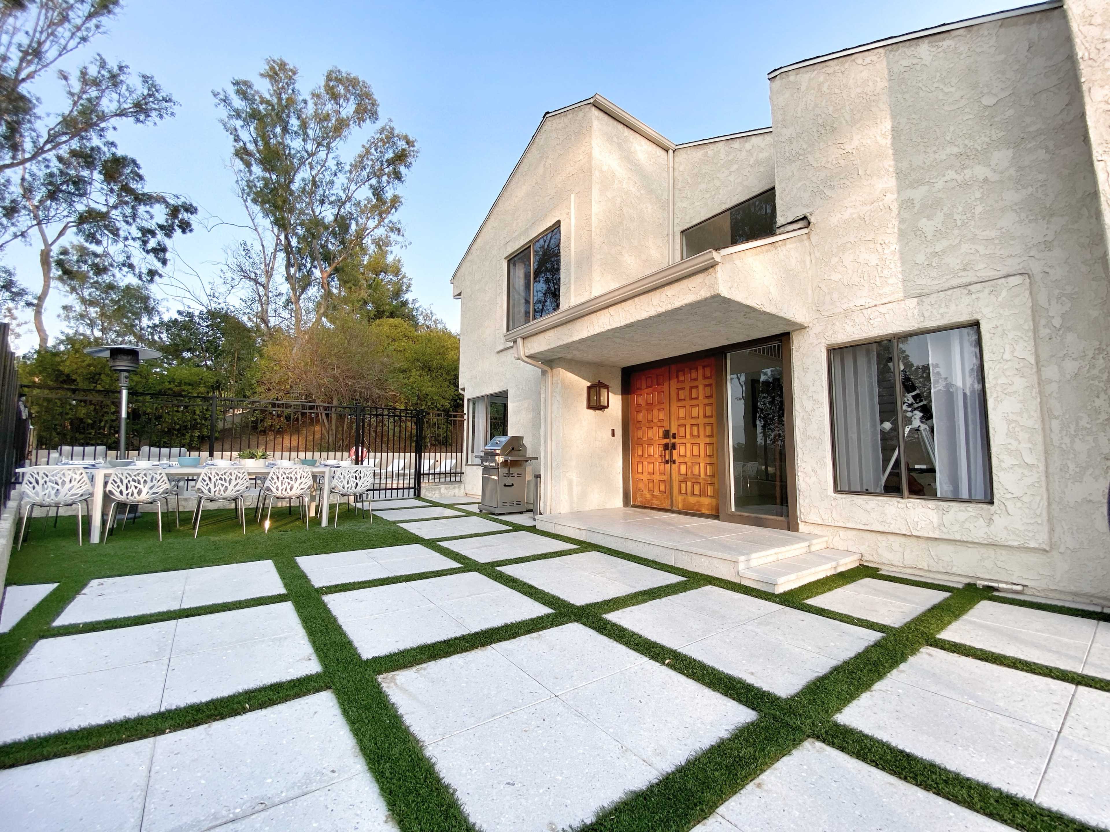 A modern house with a textured white exterior, a large wooden front door, and a landscaped patio featuring rectangular stone tiles and artificial grass.