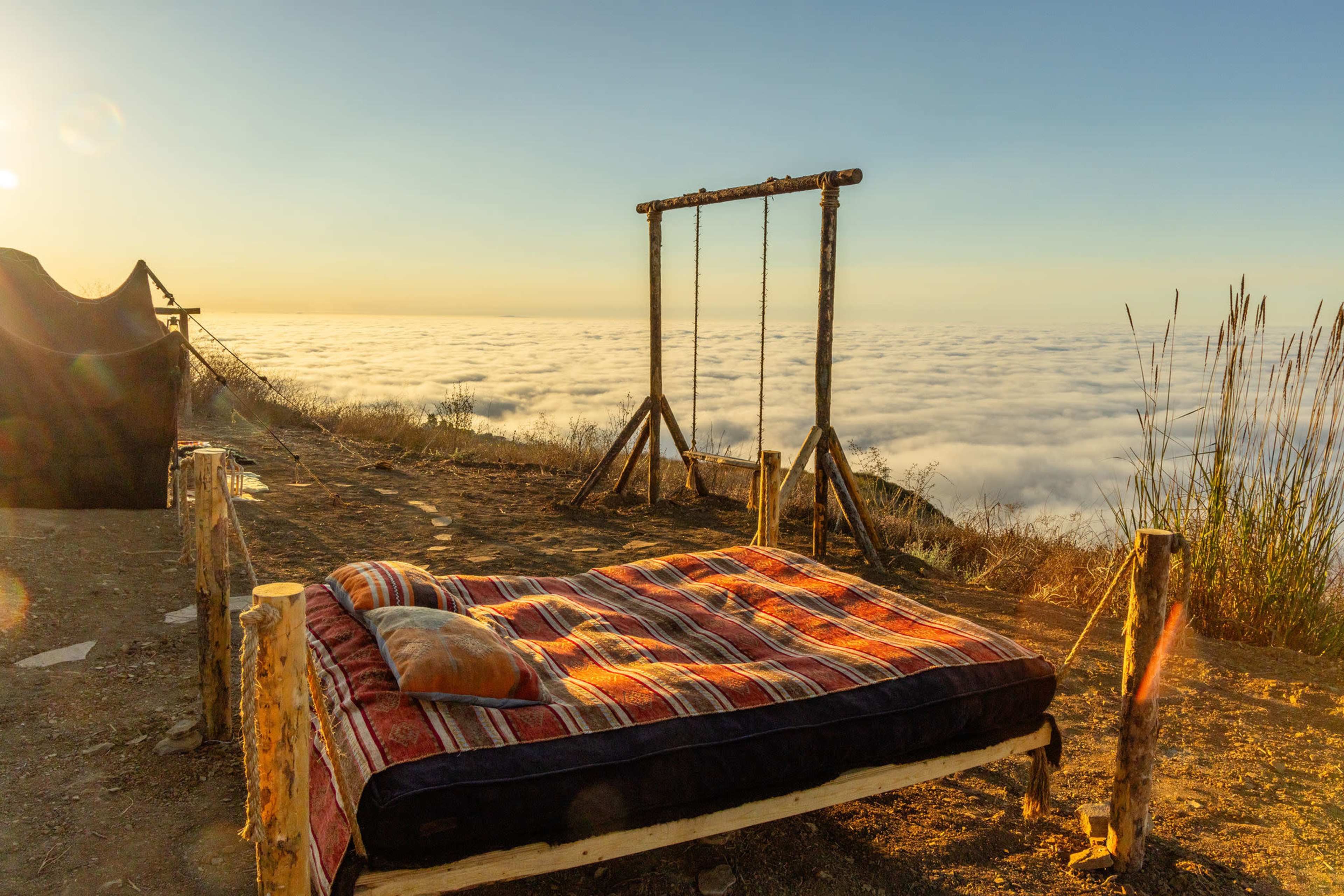 A wooden swing set stands beside a bed made of logs and blankets, overlooking a sea of clouds at sunrise.