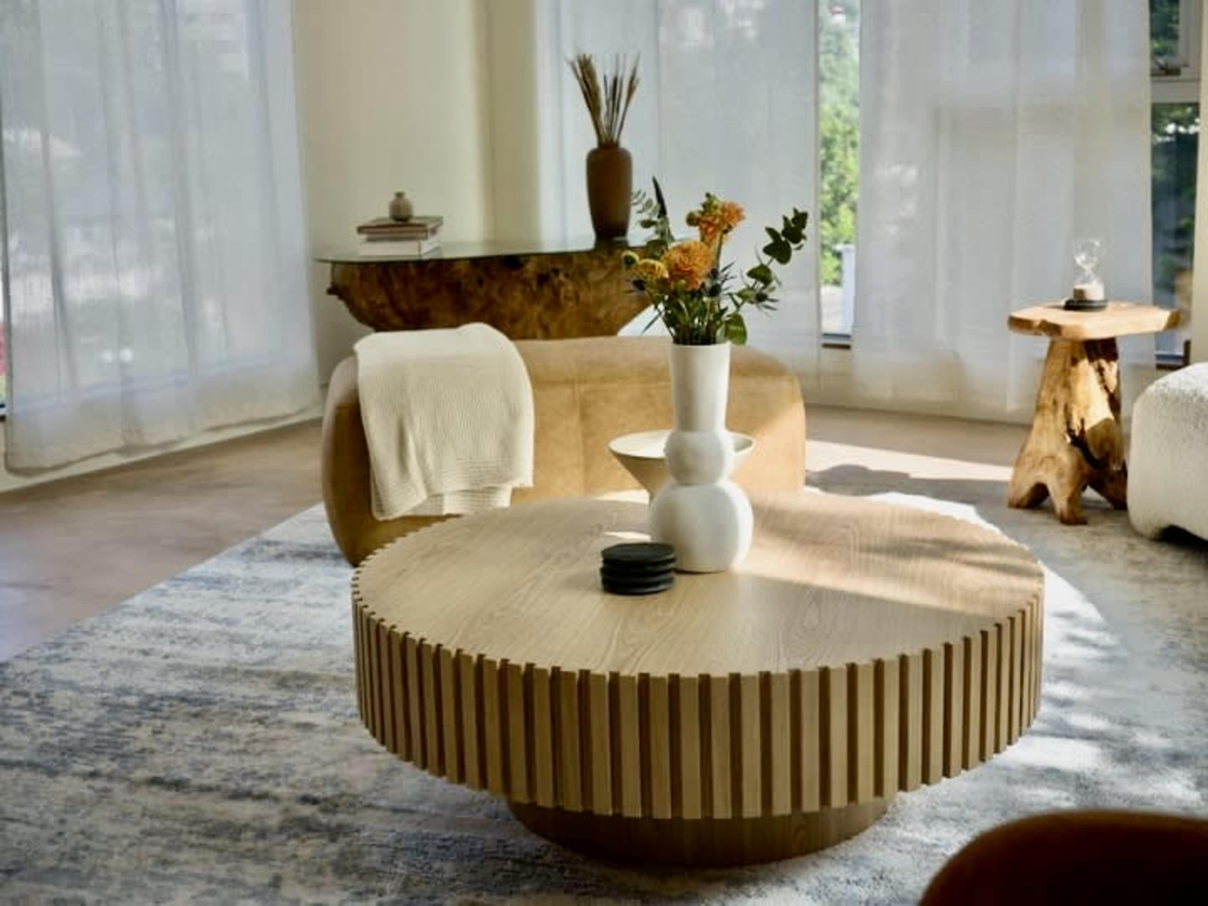 The image shows a modern living room with a round wooden coffee table, a decorative vase with flowers, and a soft, textured rug, all set against large windows covered with sheer curtains.