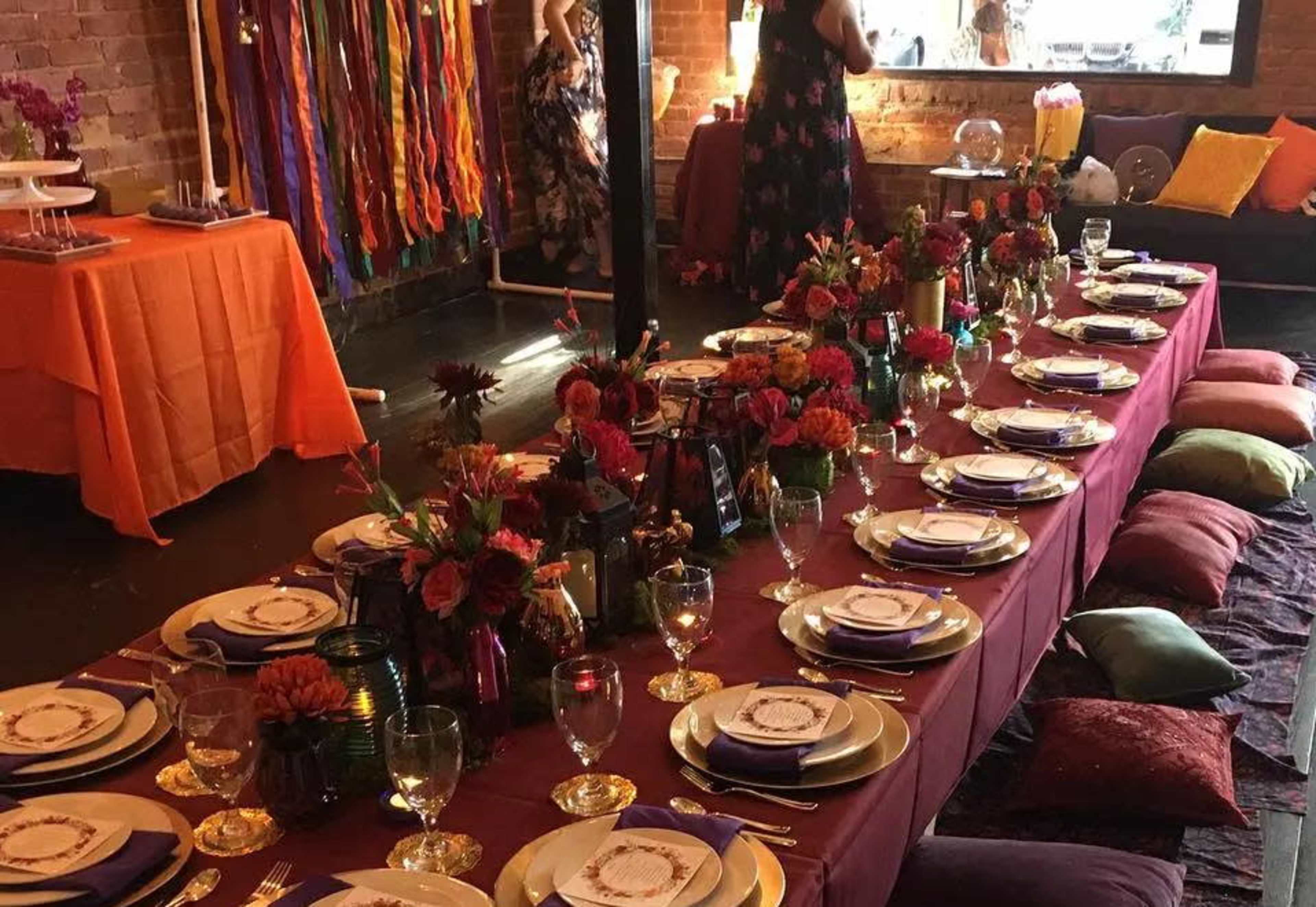 A long dining table is set with plates, glasses, and floral centerpieces, surrounded by colorful cushions in a well-decorated room.