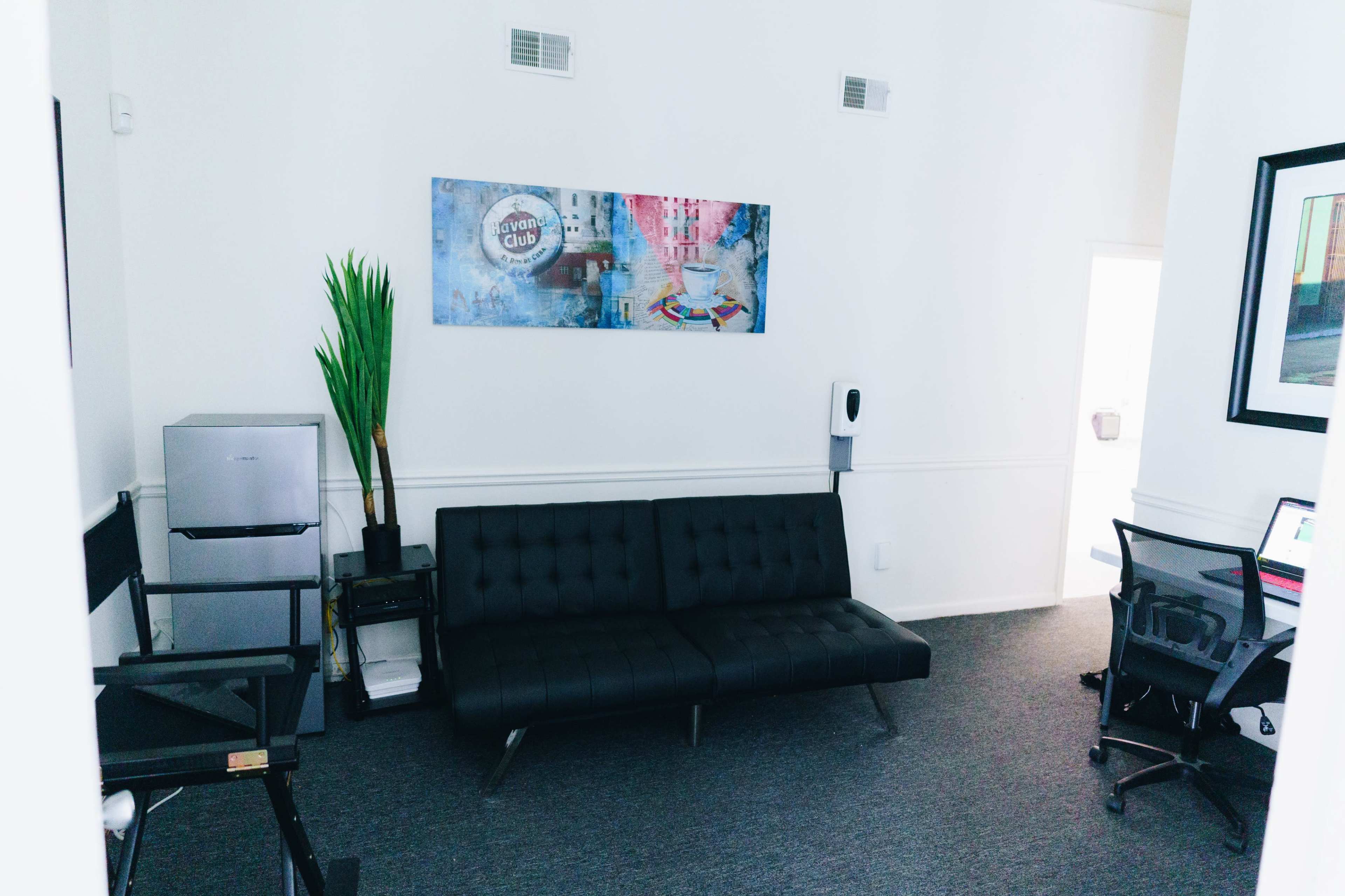 A modern office space with a black sofa, a potted plant, a refrigerator, and a desk with a computer setup against a light-colored wall.