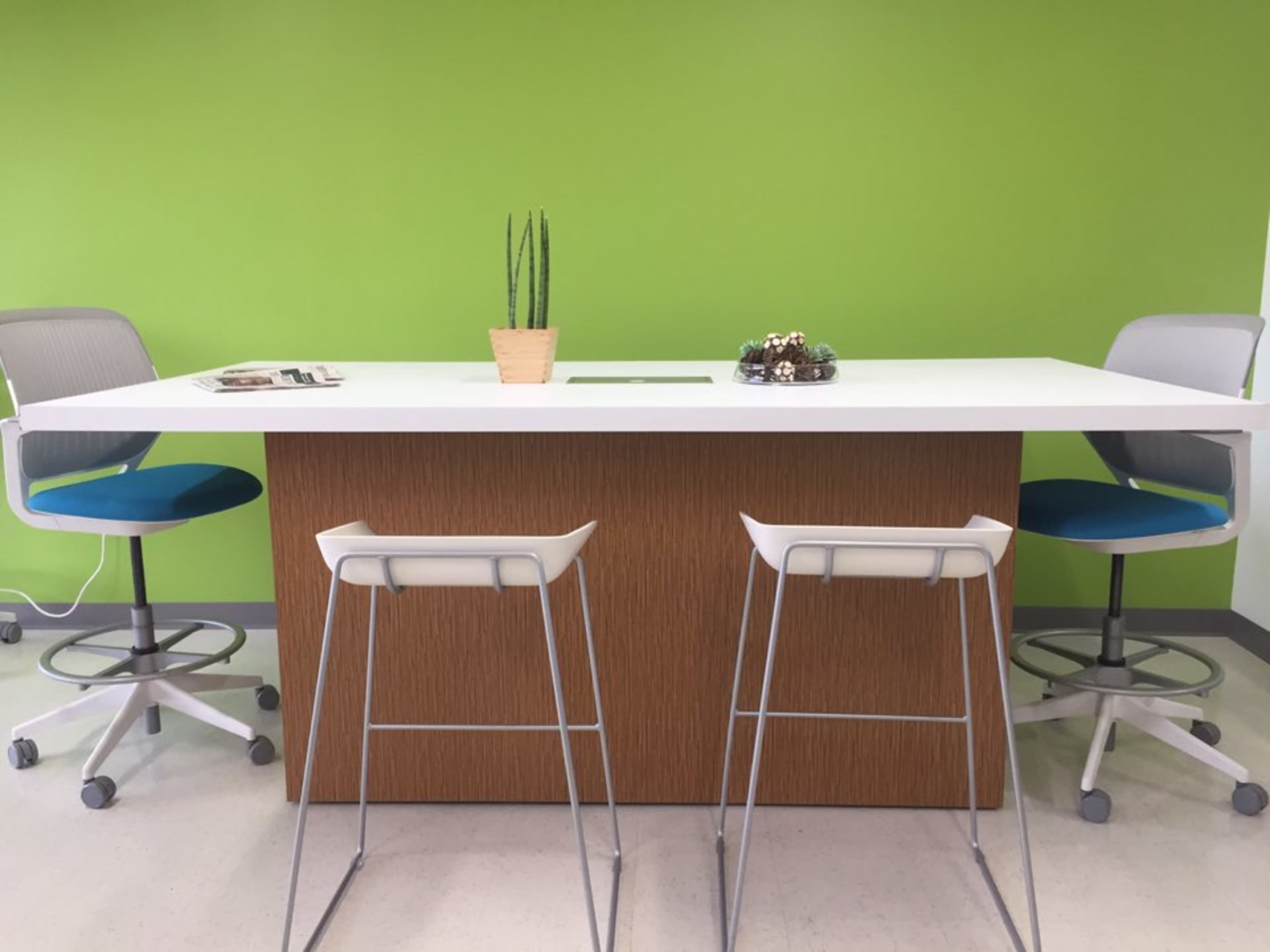 A modern workspace features a white table with a wooden base, complemented by two bar stools and a potted plant on the table.