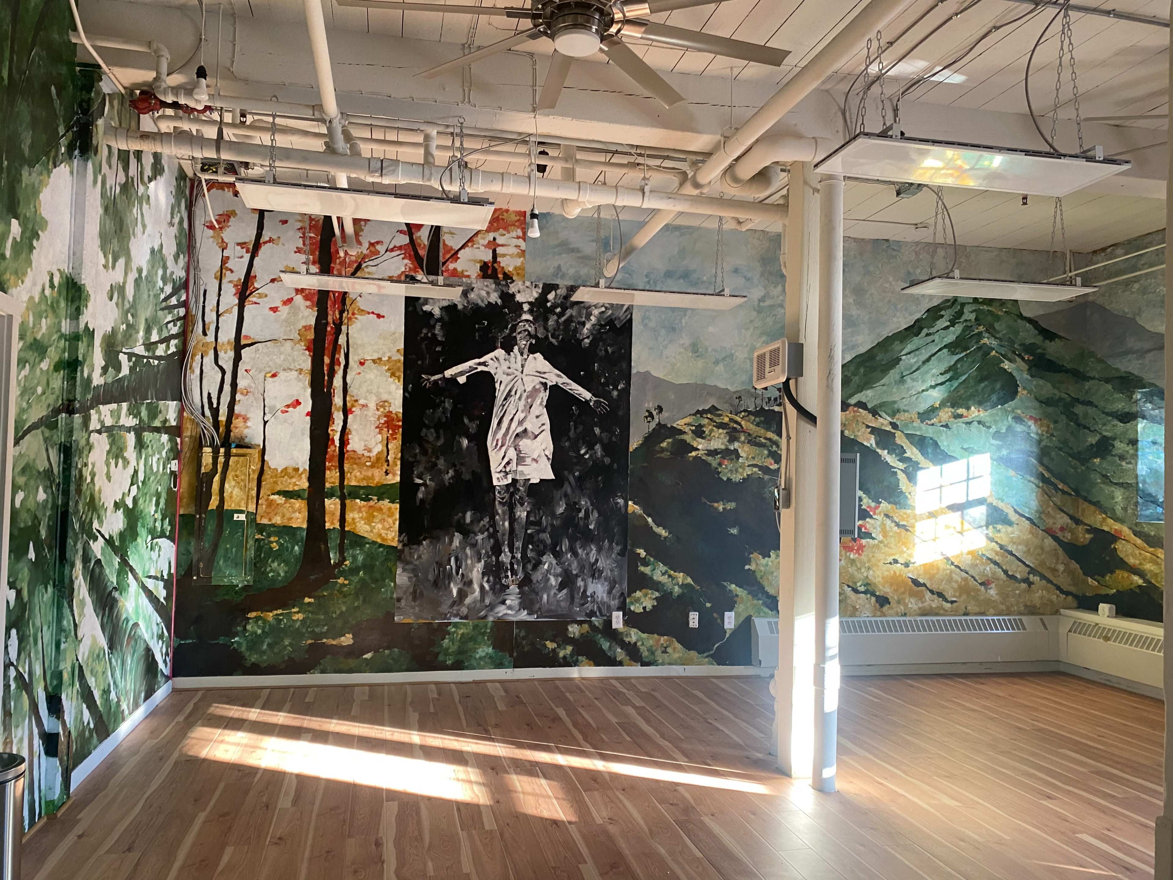An art-filled room with large murals of trees and a figure on the walls, featuring wooden flooring and natural light streaming through a window.