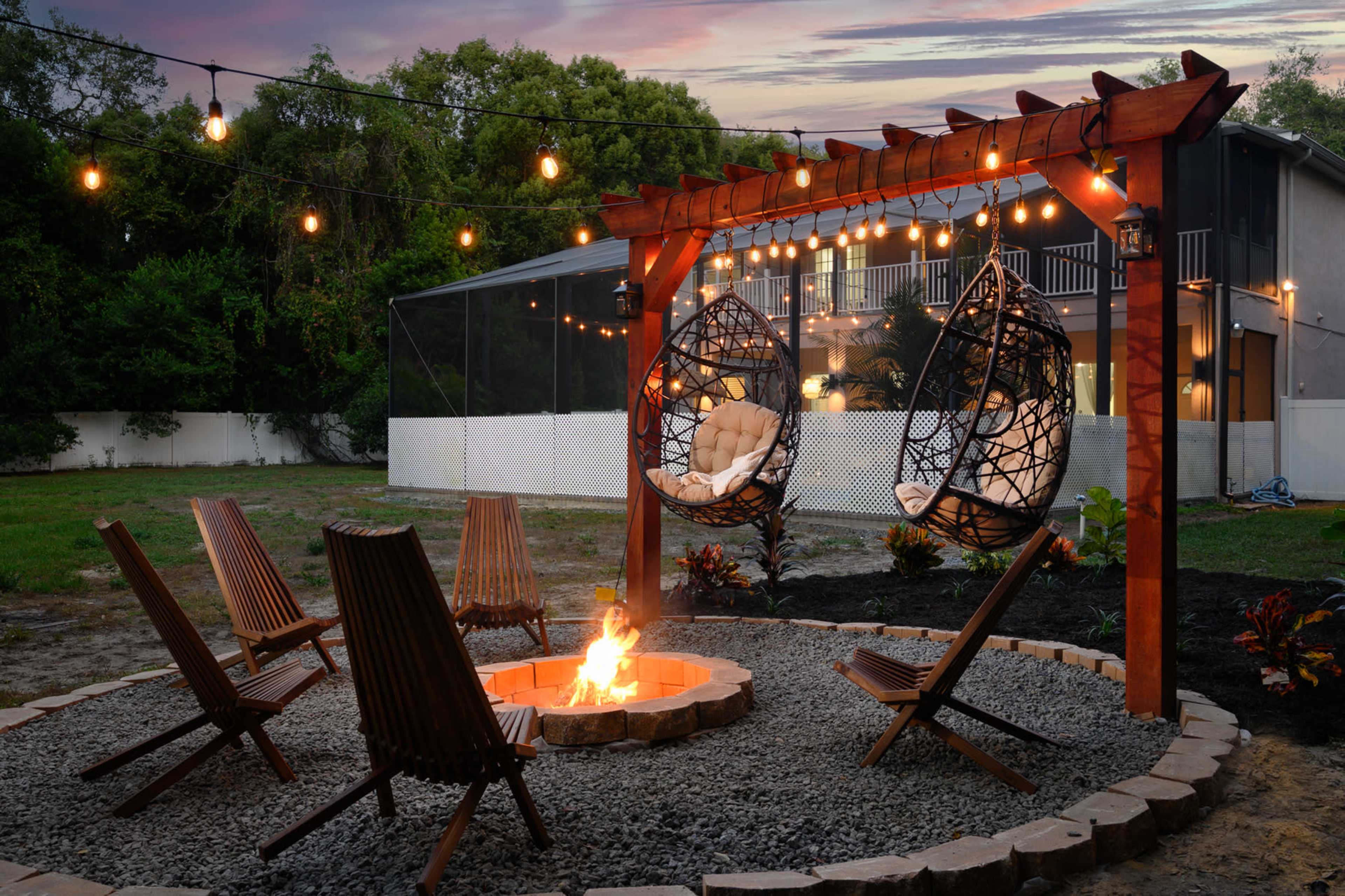 A cozy outdoor seating area features a fire pit surrounded by wooden chairs, two hanging chairs under a wooden pergola adorned with string lights.
