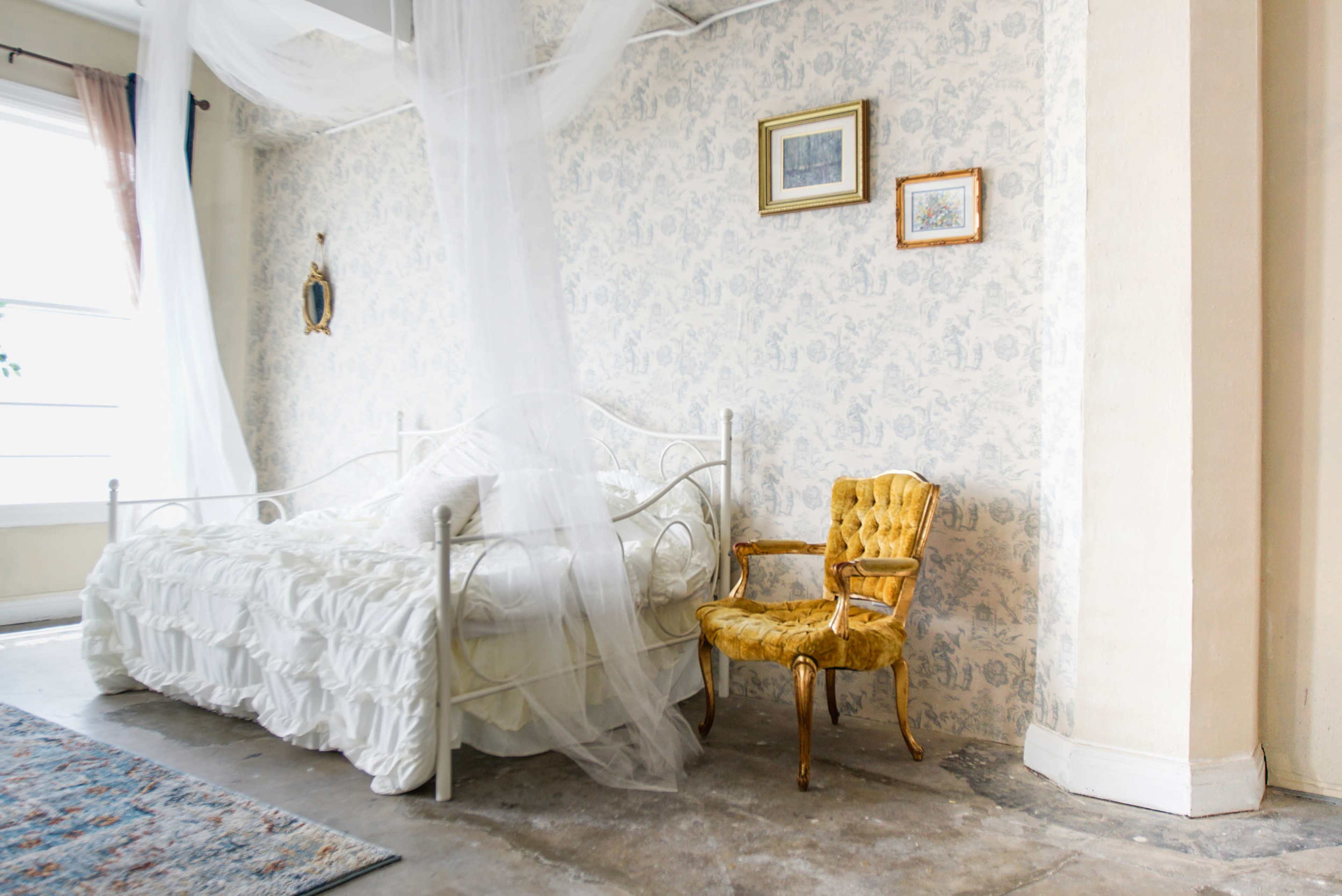 A bedroom with a white bed draped in sheer fabric and a gold chair beside it, set against a patterned wall.