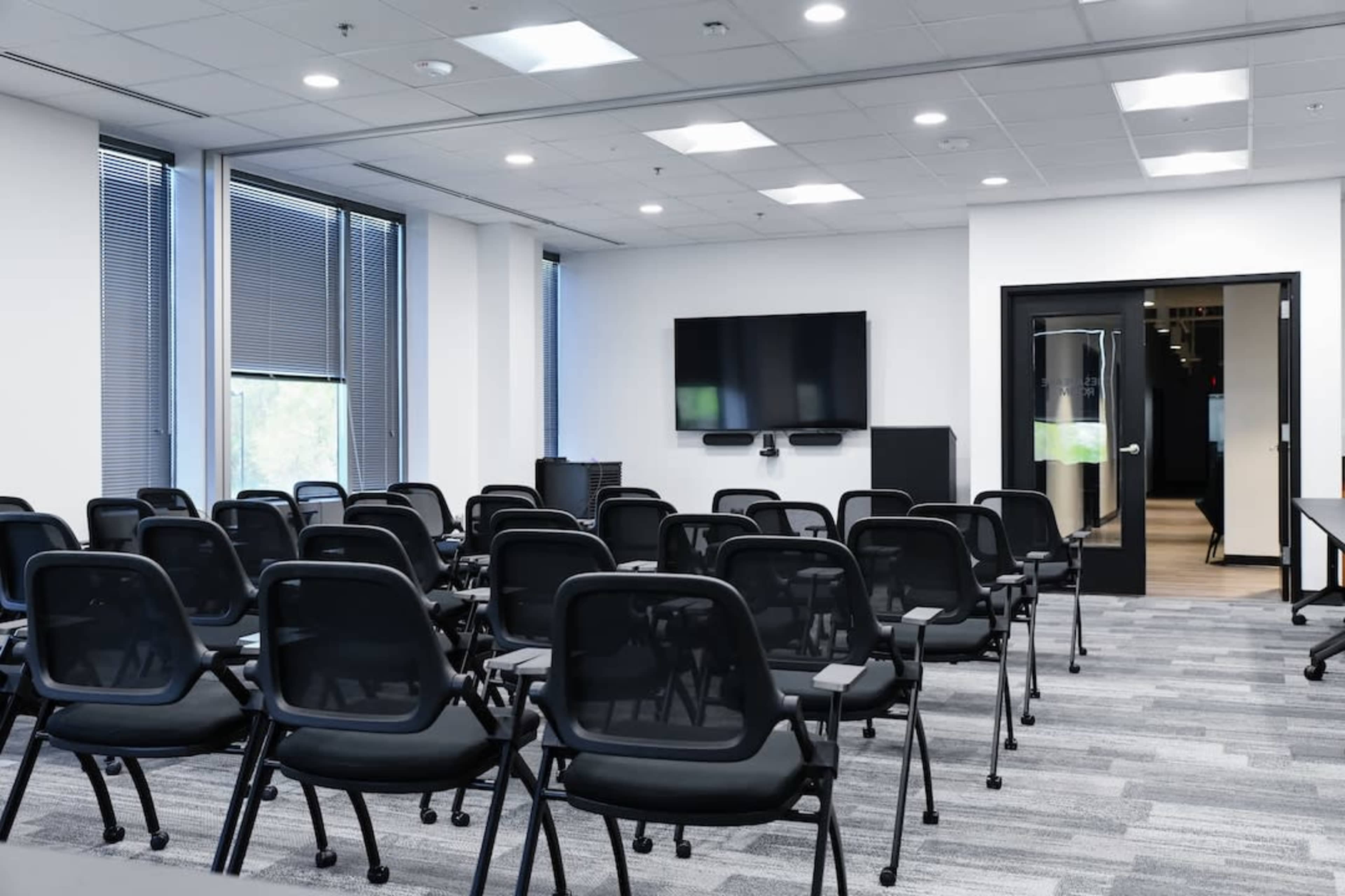 The image shows a modern conference room set up with black chairs arranged in rows facing a wall-mounted television and a door to another room.