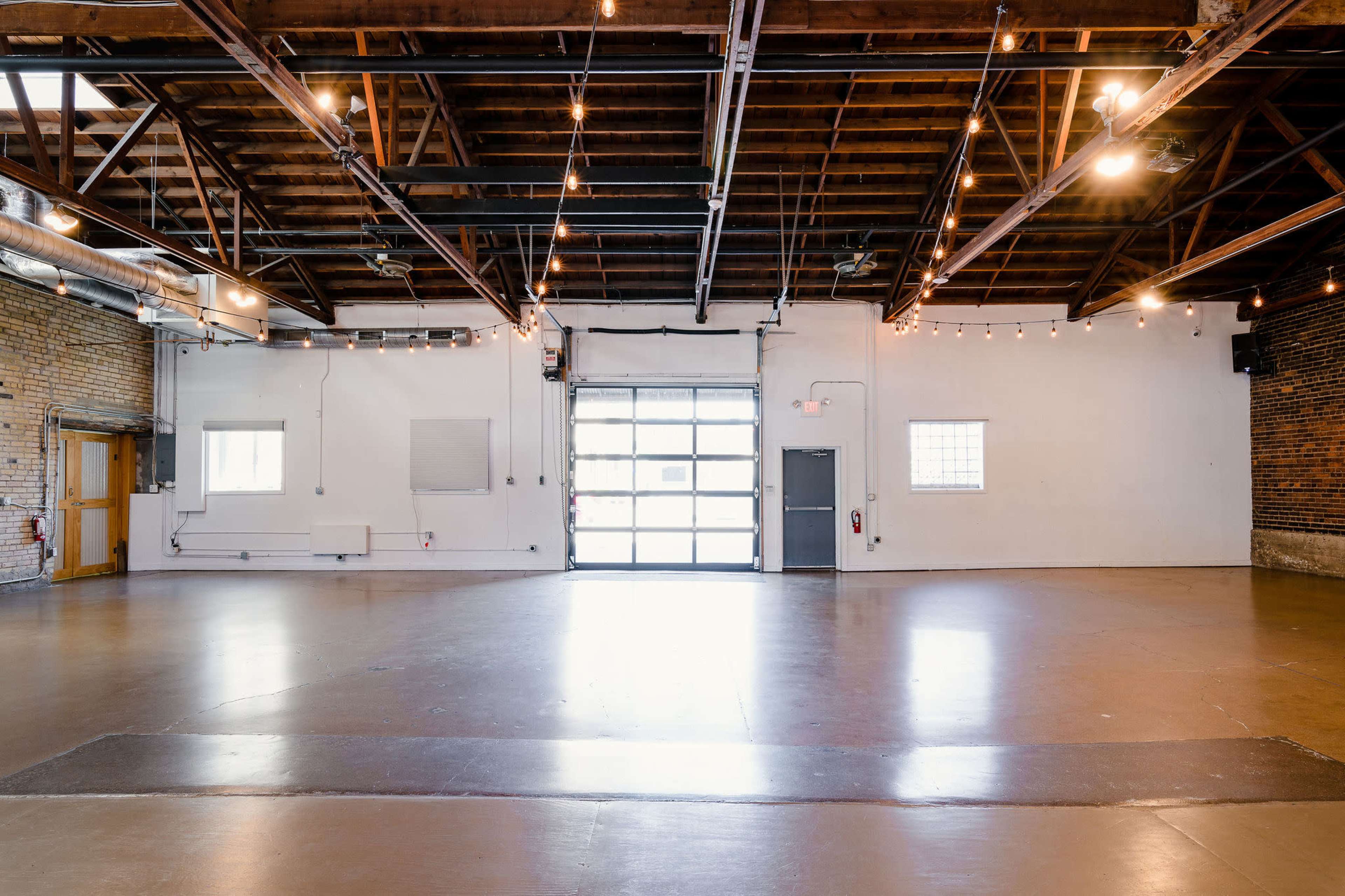 An empty industrial-style interior space with a polished concrete floor and large windows, featuring exposed wooden beams and overhead lighting.