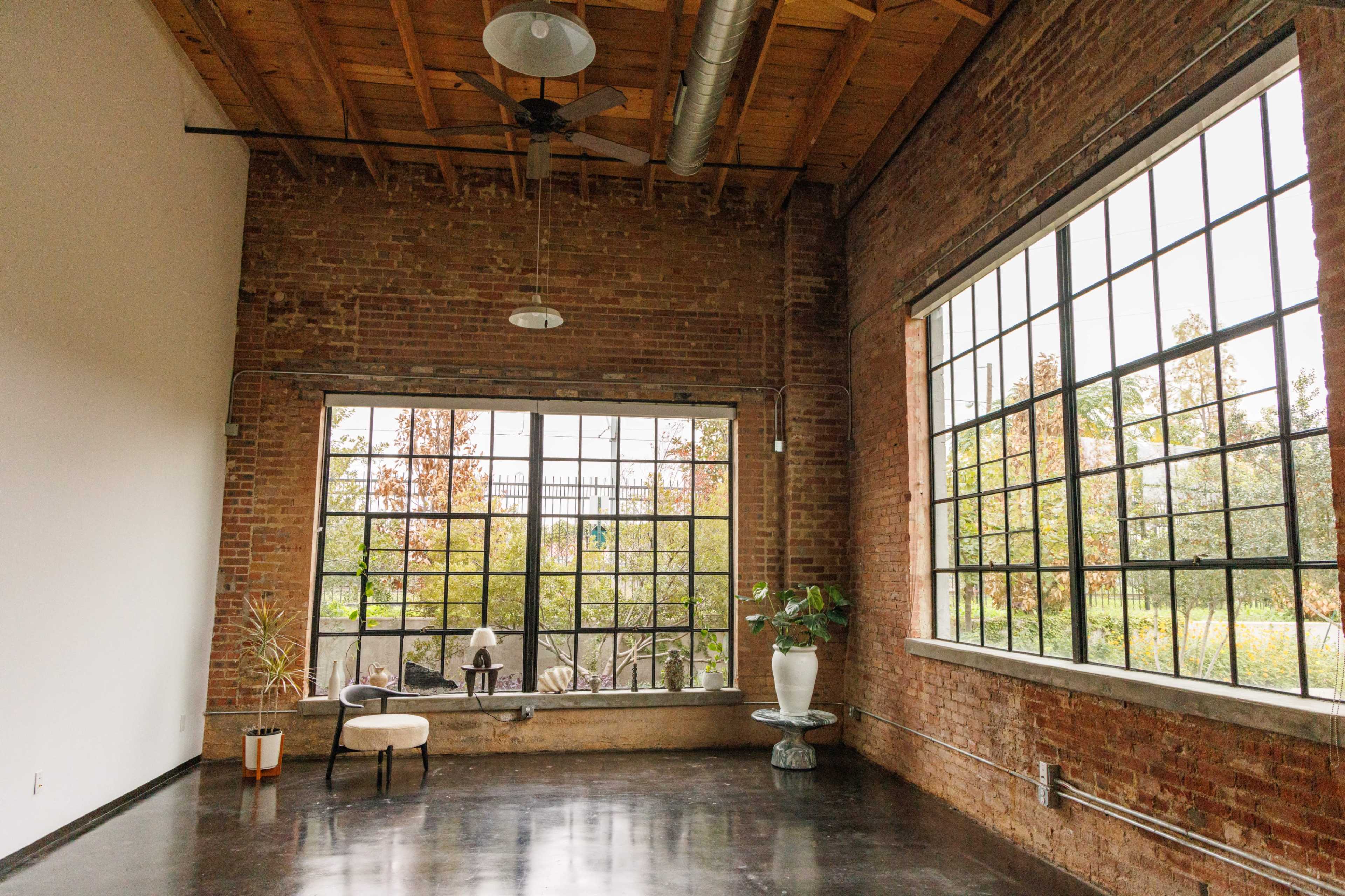 The image shows an industrial-style room with exposed brick walls, large windows, and minimal furnishings, including two chairs and potted plants.