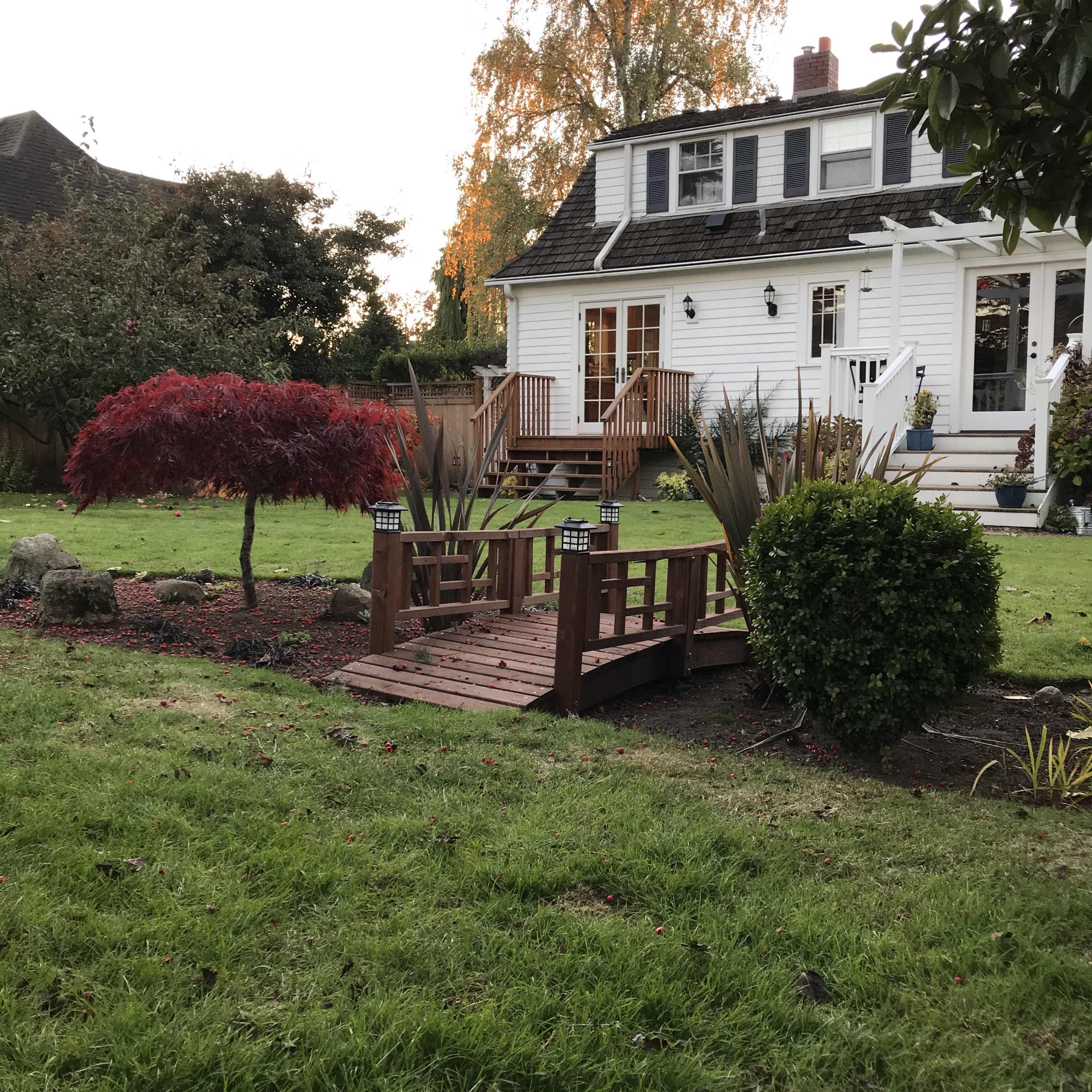 A wooden bridge crosses over a small garden area with decorative plants and a red Japanese maple in front of a white house.