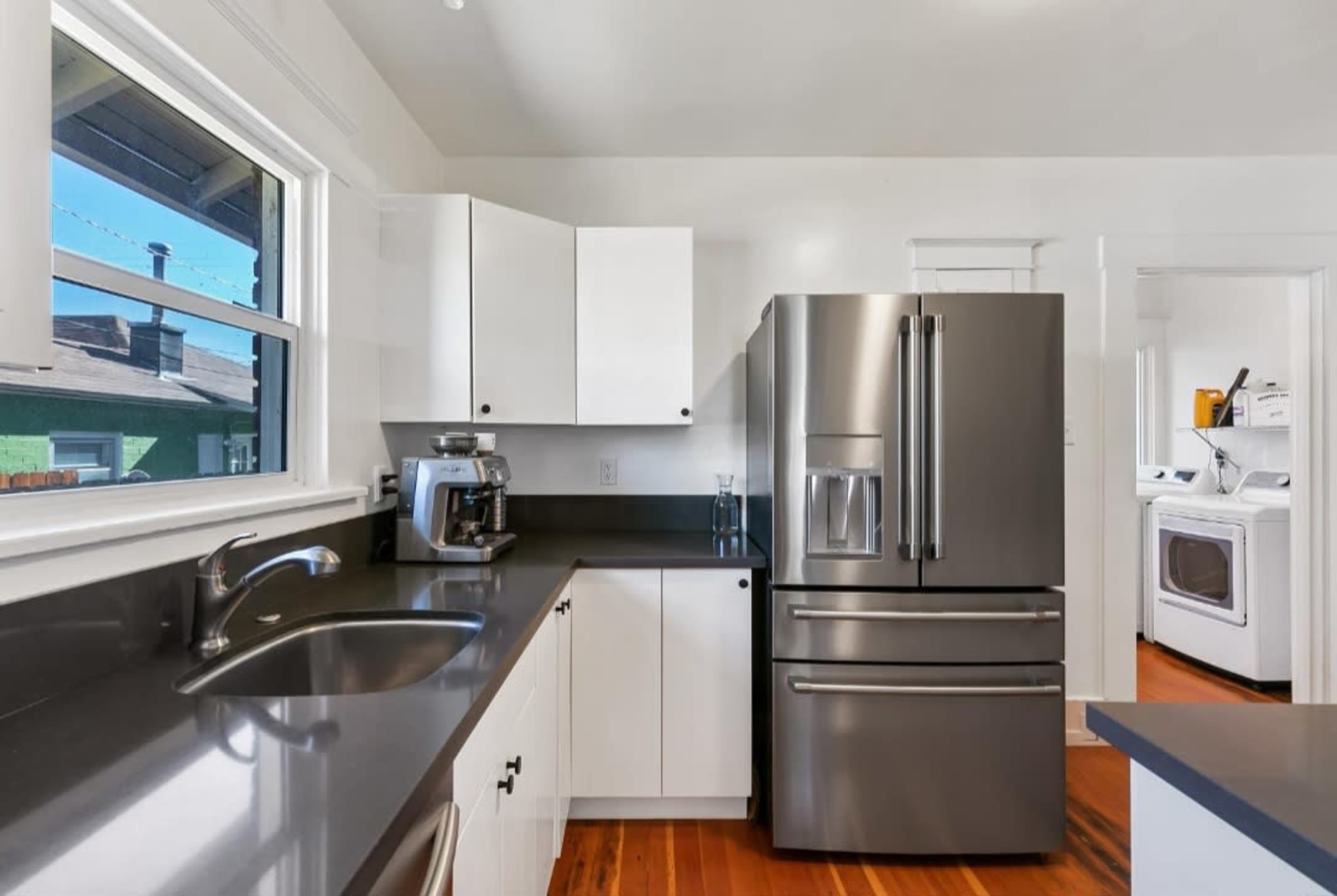 A modern kitchen features a stainless steel refrigerator, black countertop, and white cabinets, with a view into a laundry area through an adjacent doorway.