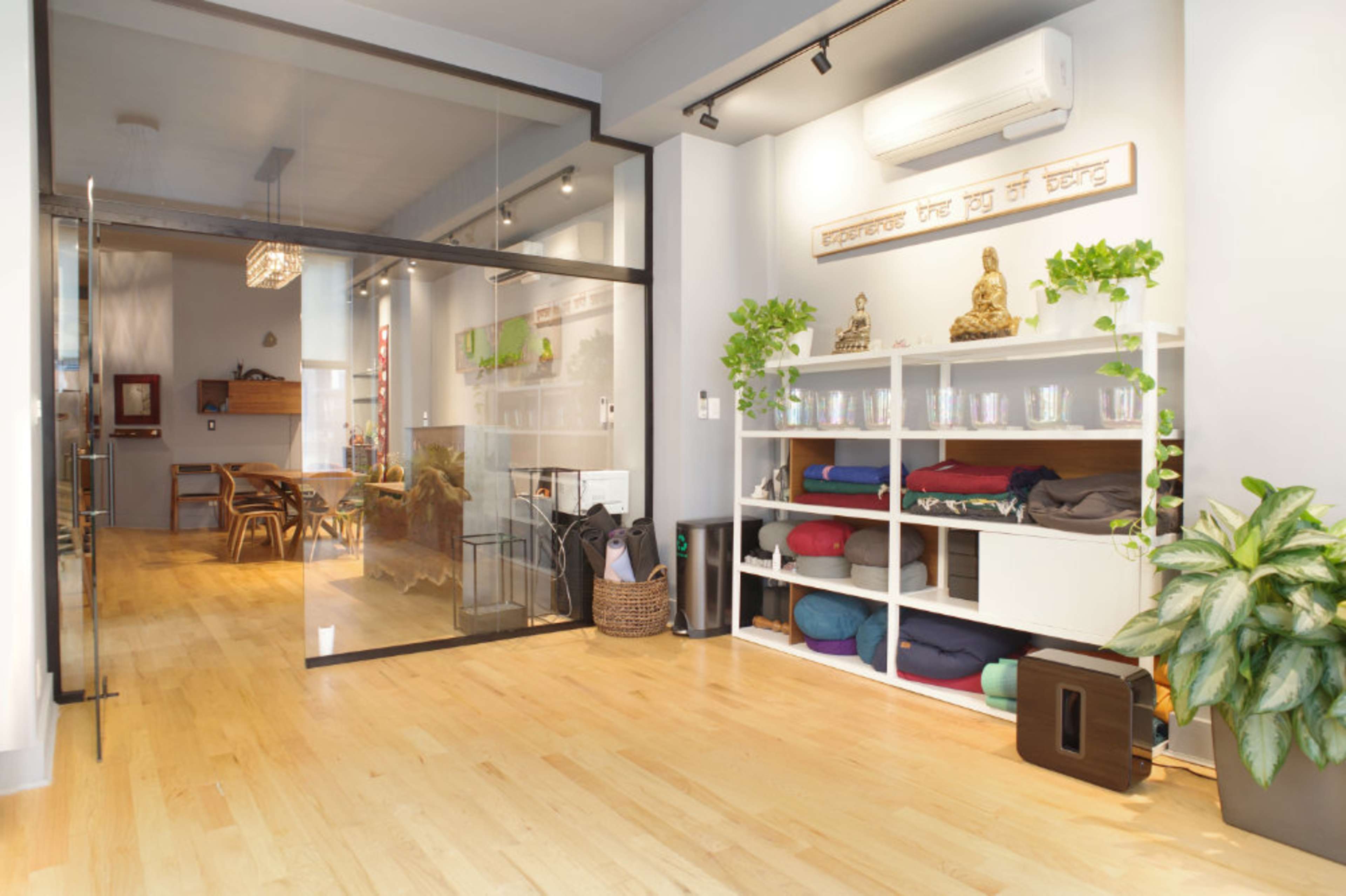 The image shows a modern, minimalist interior space with wooden flooring, glass partitions, and shelves filled with yoga mats and cushions, alongside plants and decorative elements.