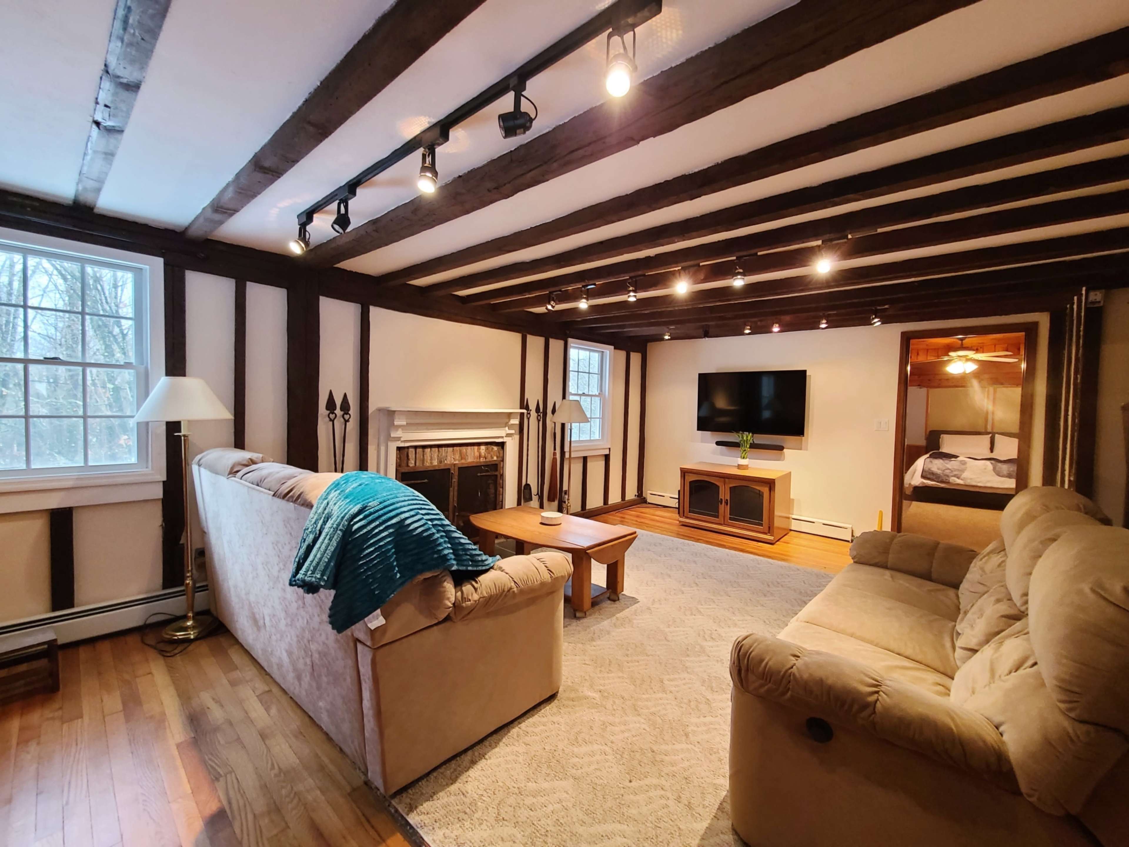 A living room with wooden beams, a fireplace, a flat-screen TV mounted on the wall, and a doorway leading to a bedroom.