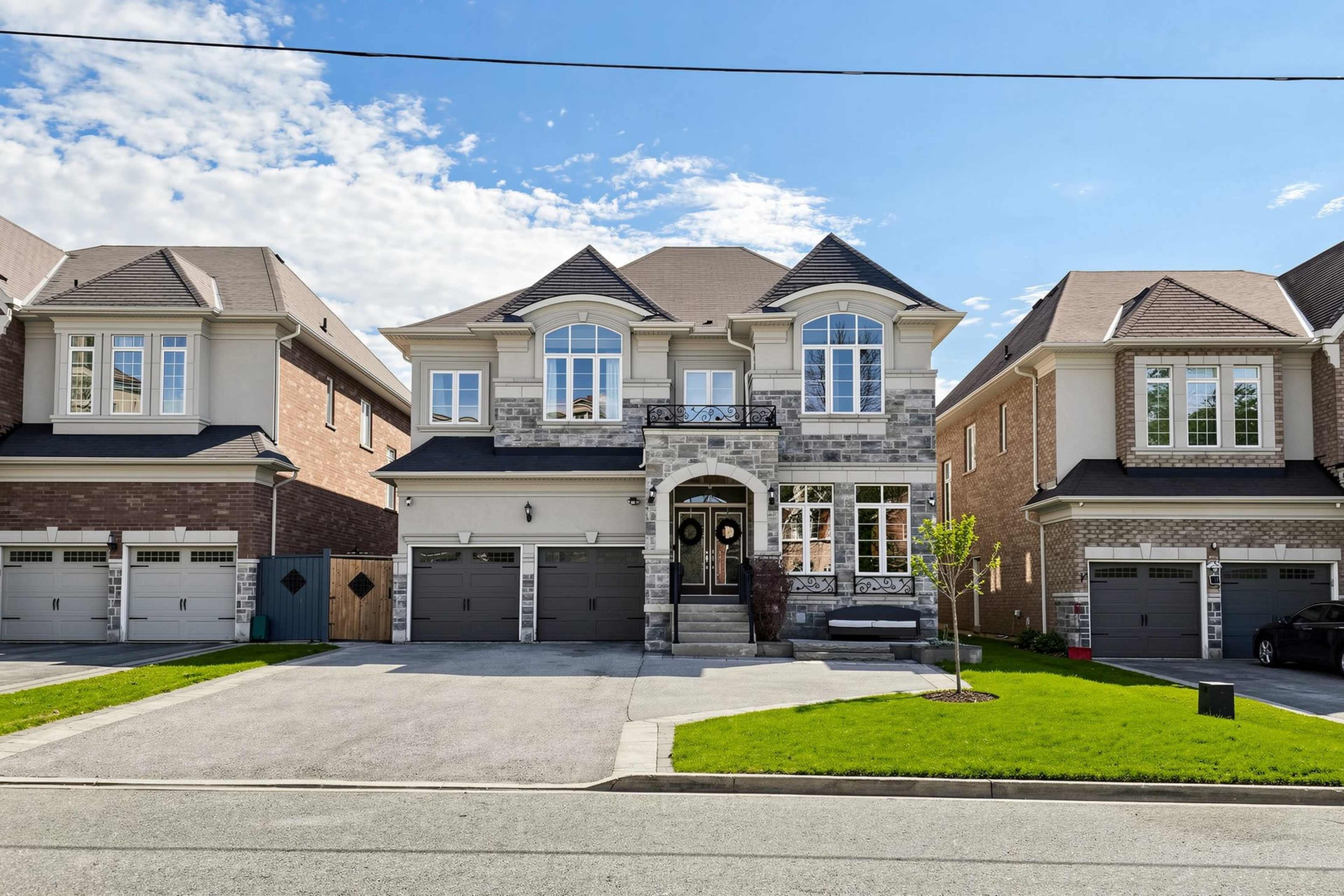 The image shows a large, two-story house with a stone and brick exterior, flanked by similar homes, and a neatly landscaped front yard with a driveway.
