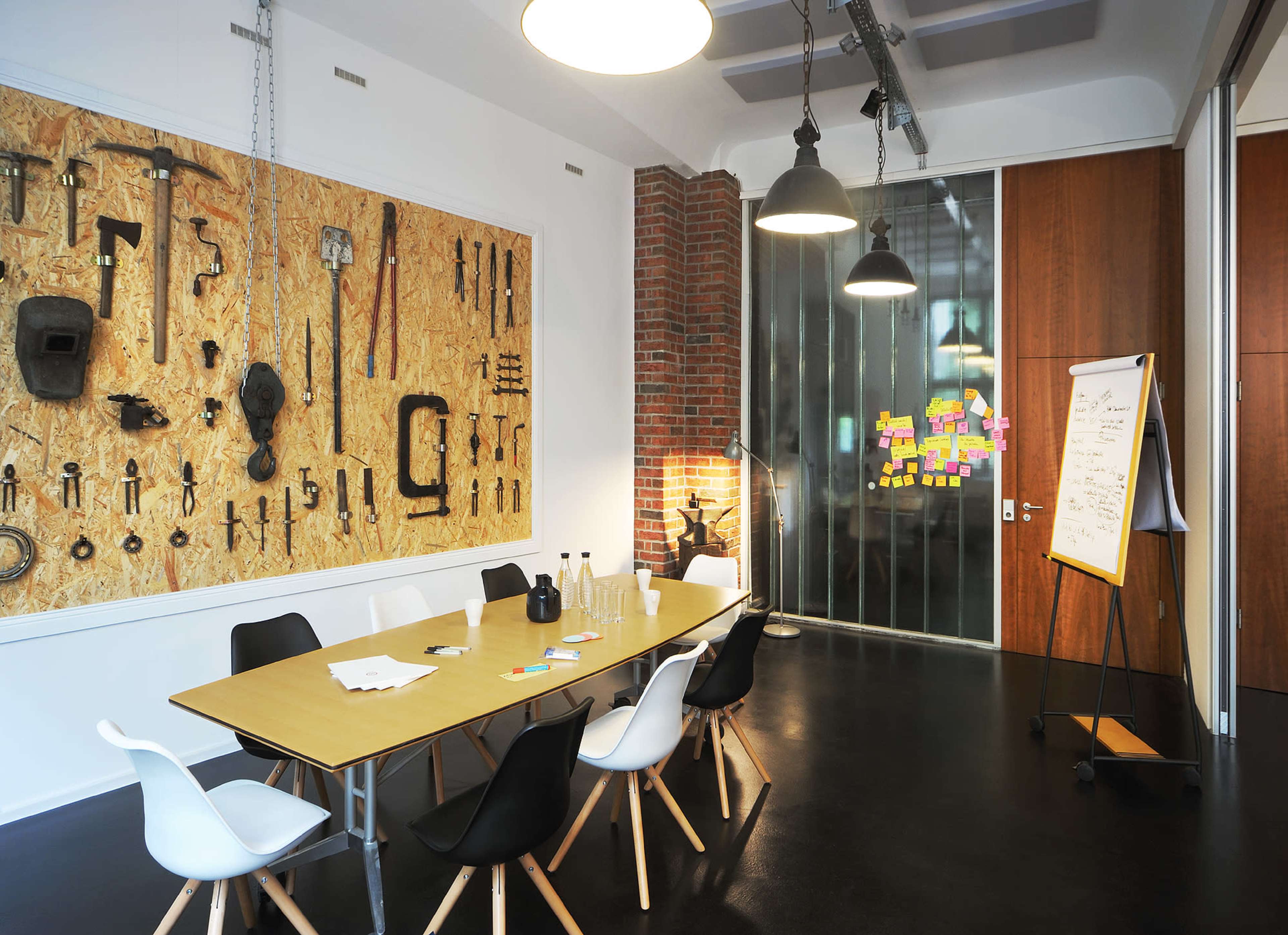 A modern meeting room features a central wooden table surrounded by black and white chairs, with a wall adorned by various tools and a glass partition displaying colorful notes.