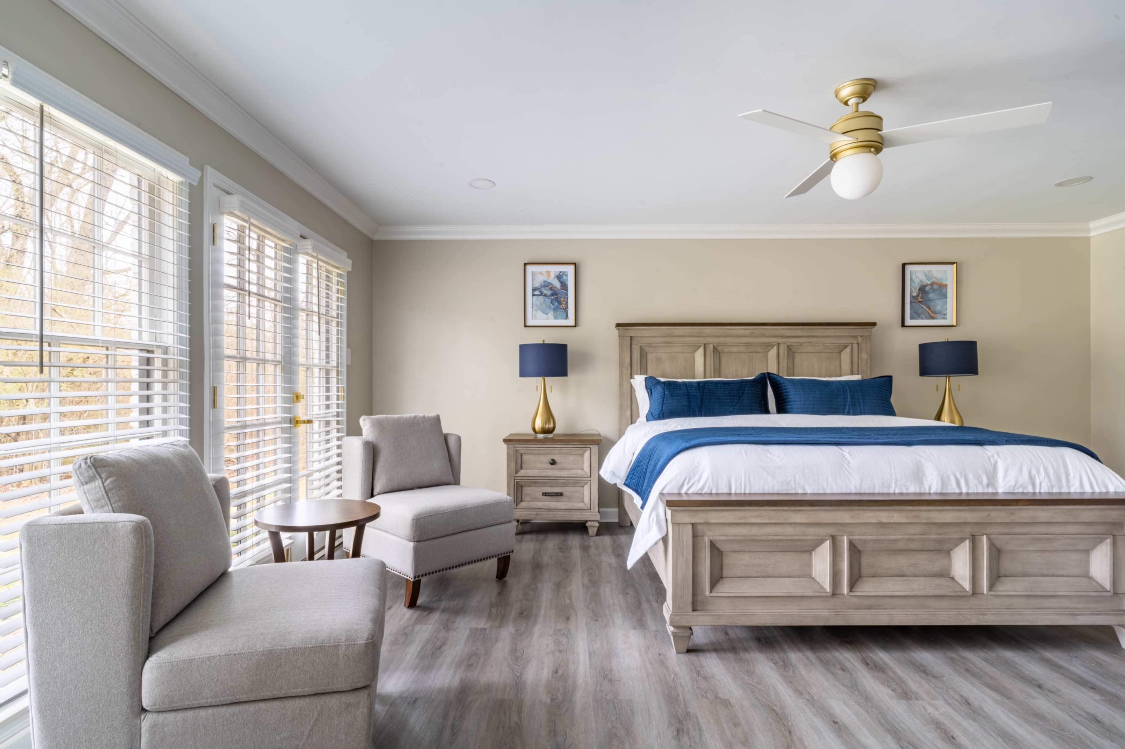 The image shows a well-furnished bedroom featuring a large bed with blue bedding, two bedside lamps, a small wooden table, and a seating area with a chair and a side table near a window with blinds.