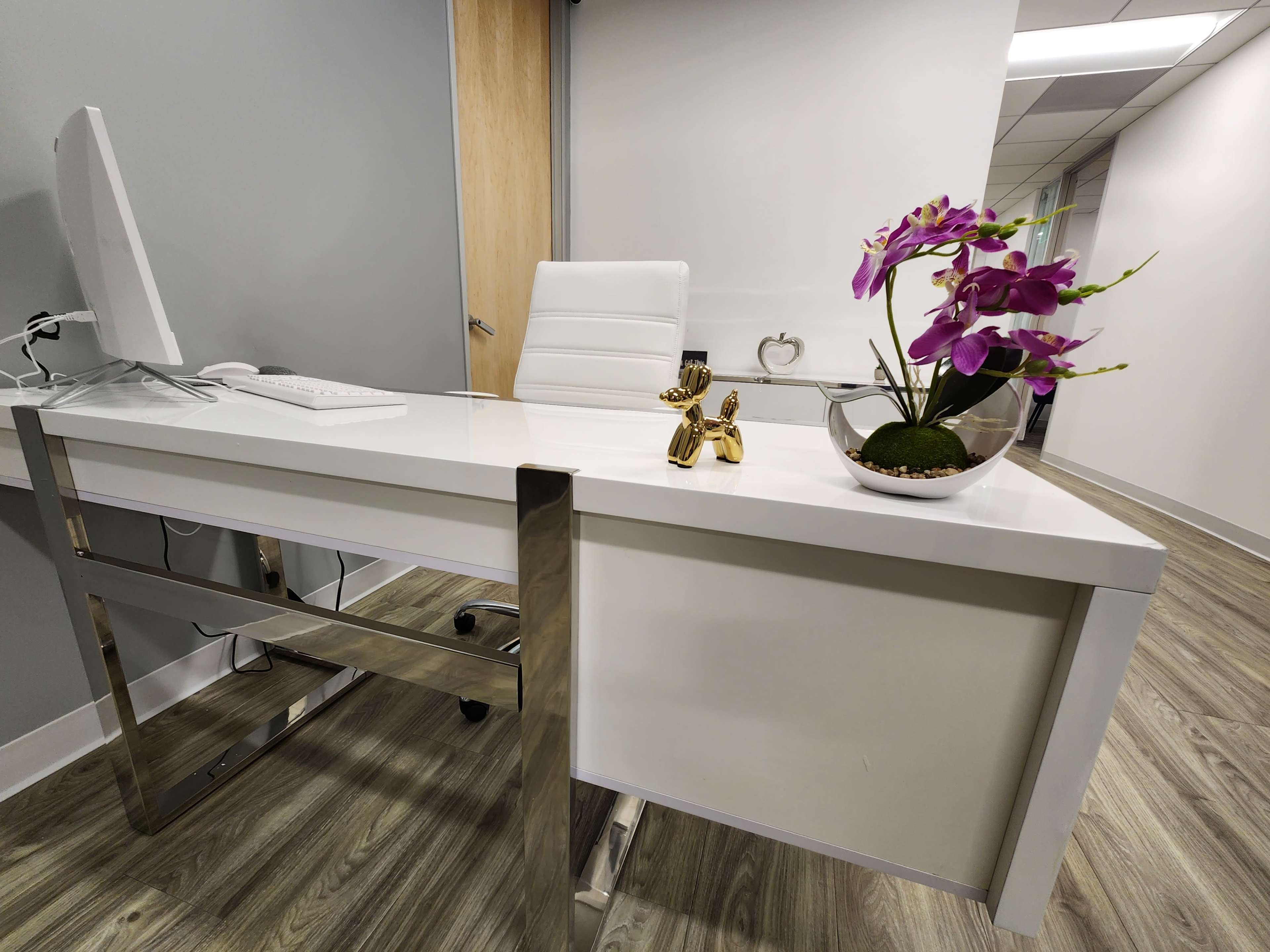 The image depicts a modern office space featuring a sleek white desk with a computer, a chair, and a decorative vase with purple orchids.