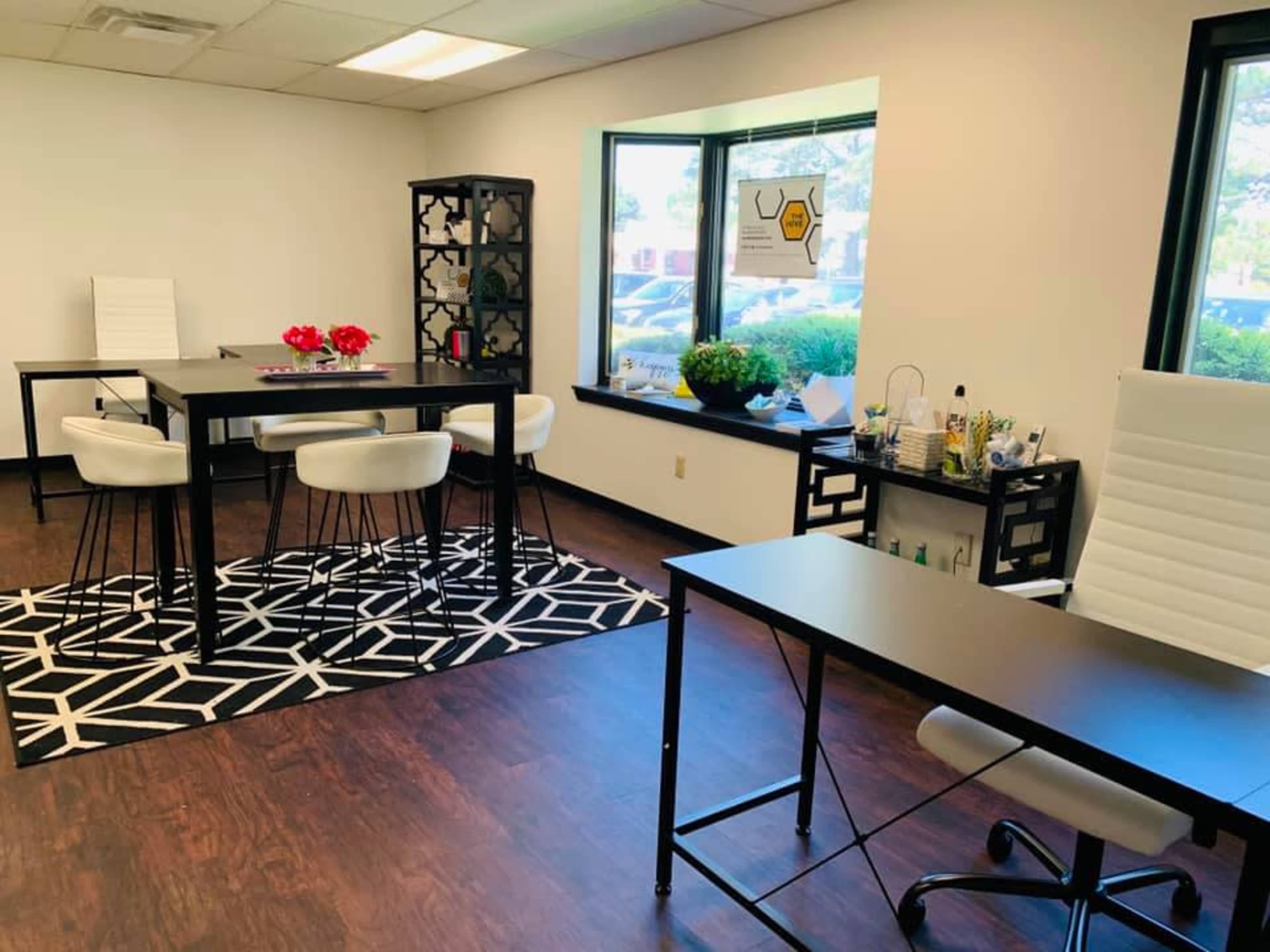A modern office space with two black desks, white chairs, a patterned rug, and a large window letting in natural light.