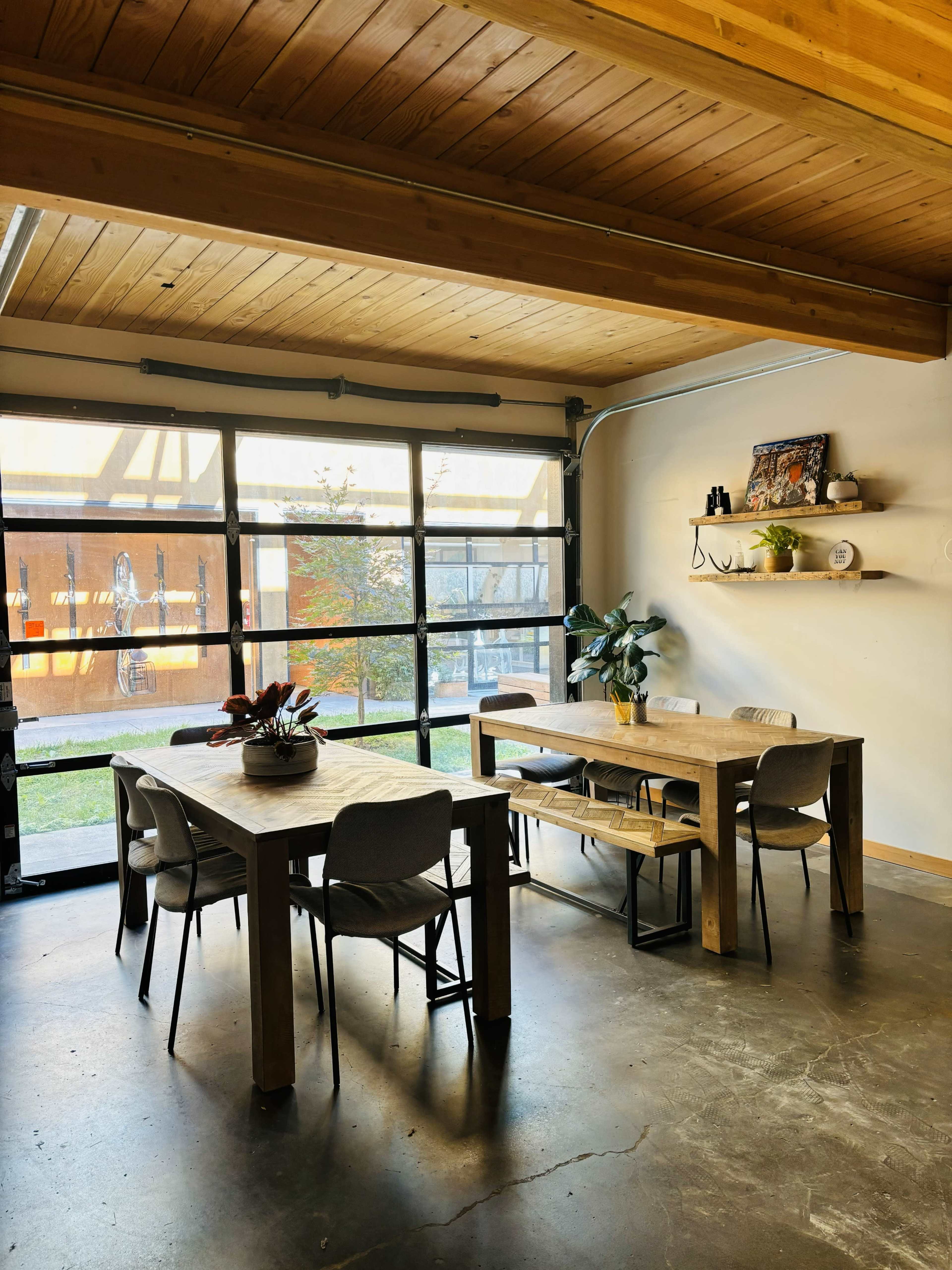 The image shows a modern dining area with a large wooden table, several chairs, and a garage-style door that opens to an outdoor space.