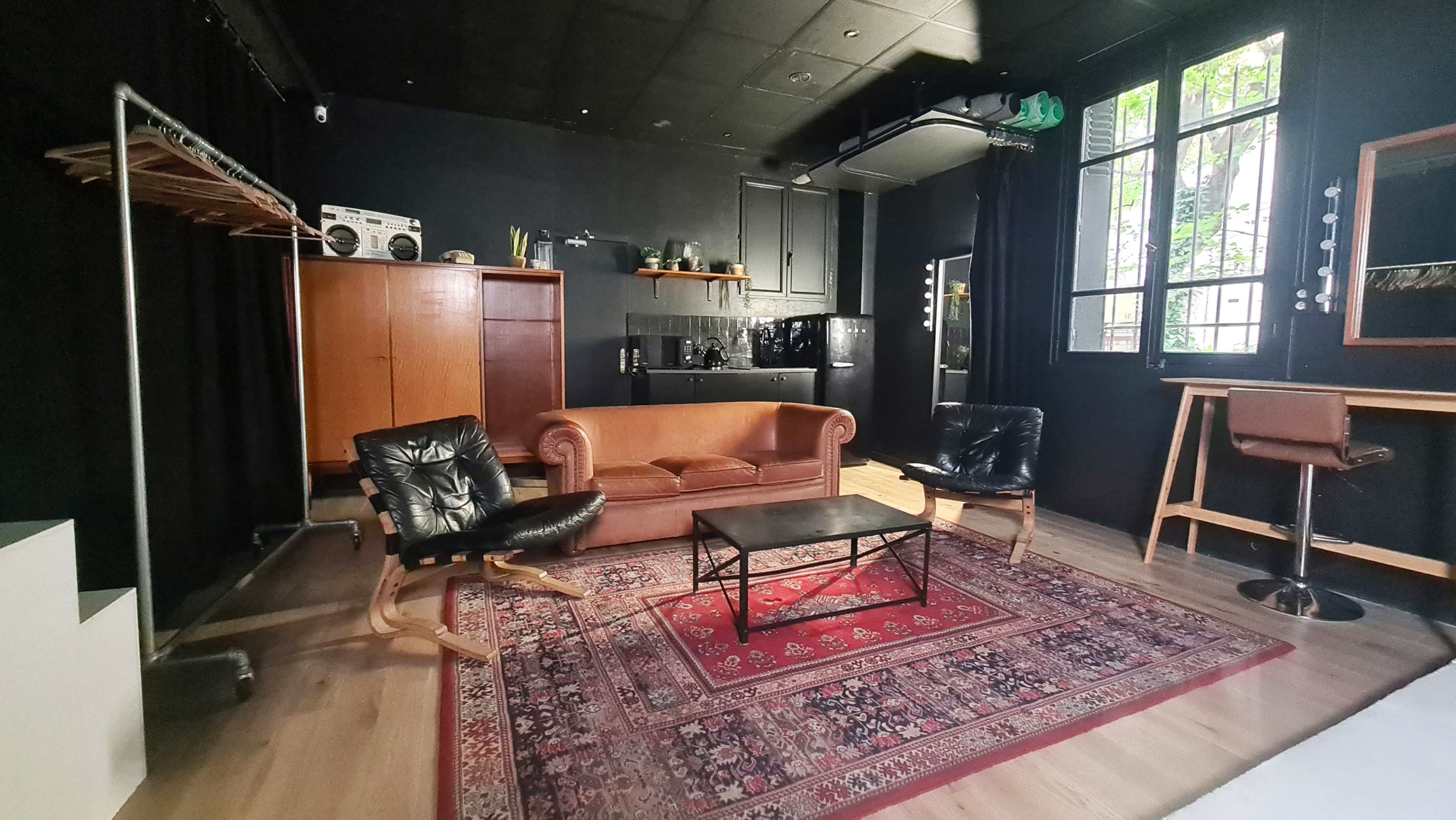 The image shows a stylish, black-walled room featuring a brown leather sofa, two black armchairs, a wooden coffee table on a patterned rug, and a bar area with high stools.