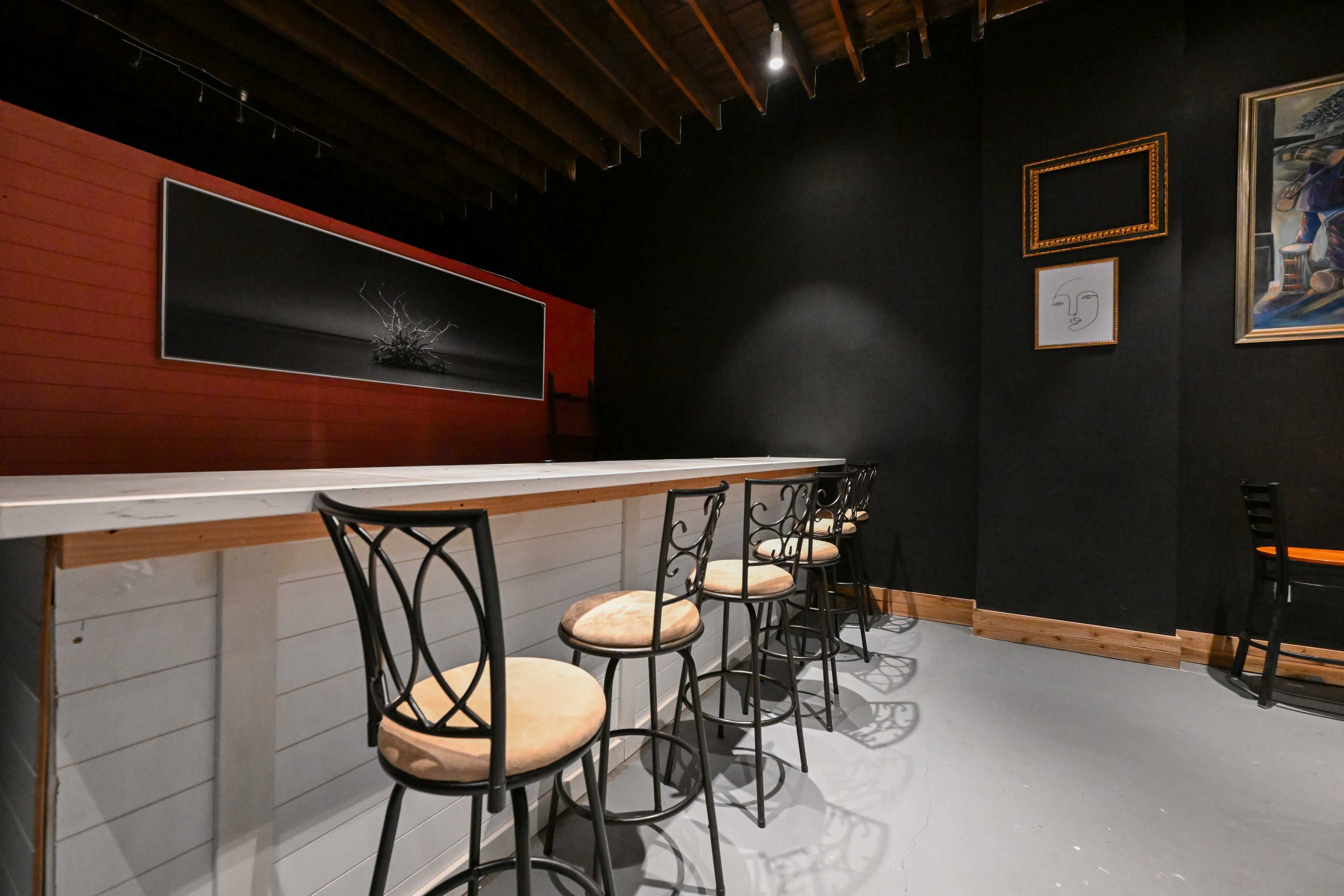 The image shows an empty bar area with a long white countertop and several black chairs against dark walls, featuring minimal artwork.