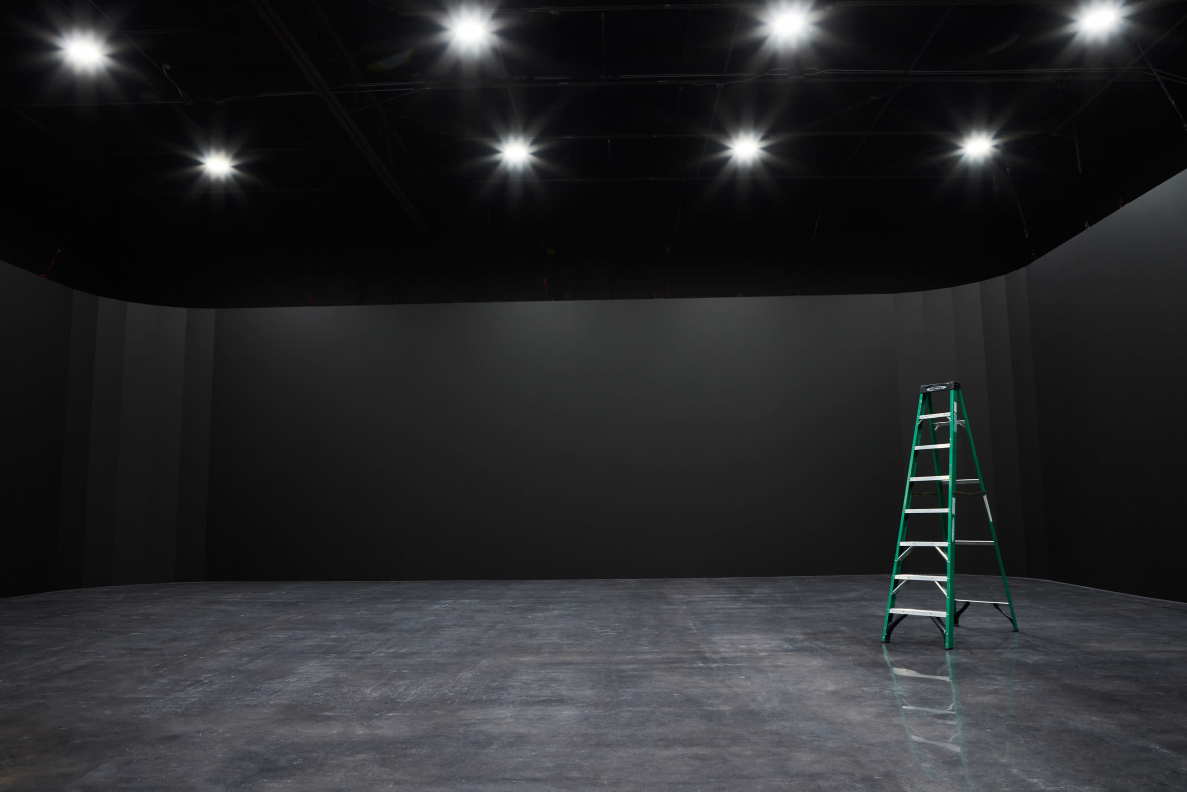 A green ladder stands against a blank black wall in an empty room with polished concrete flooring, illuminated by overhead lights.
