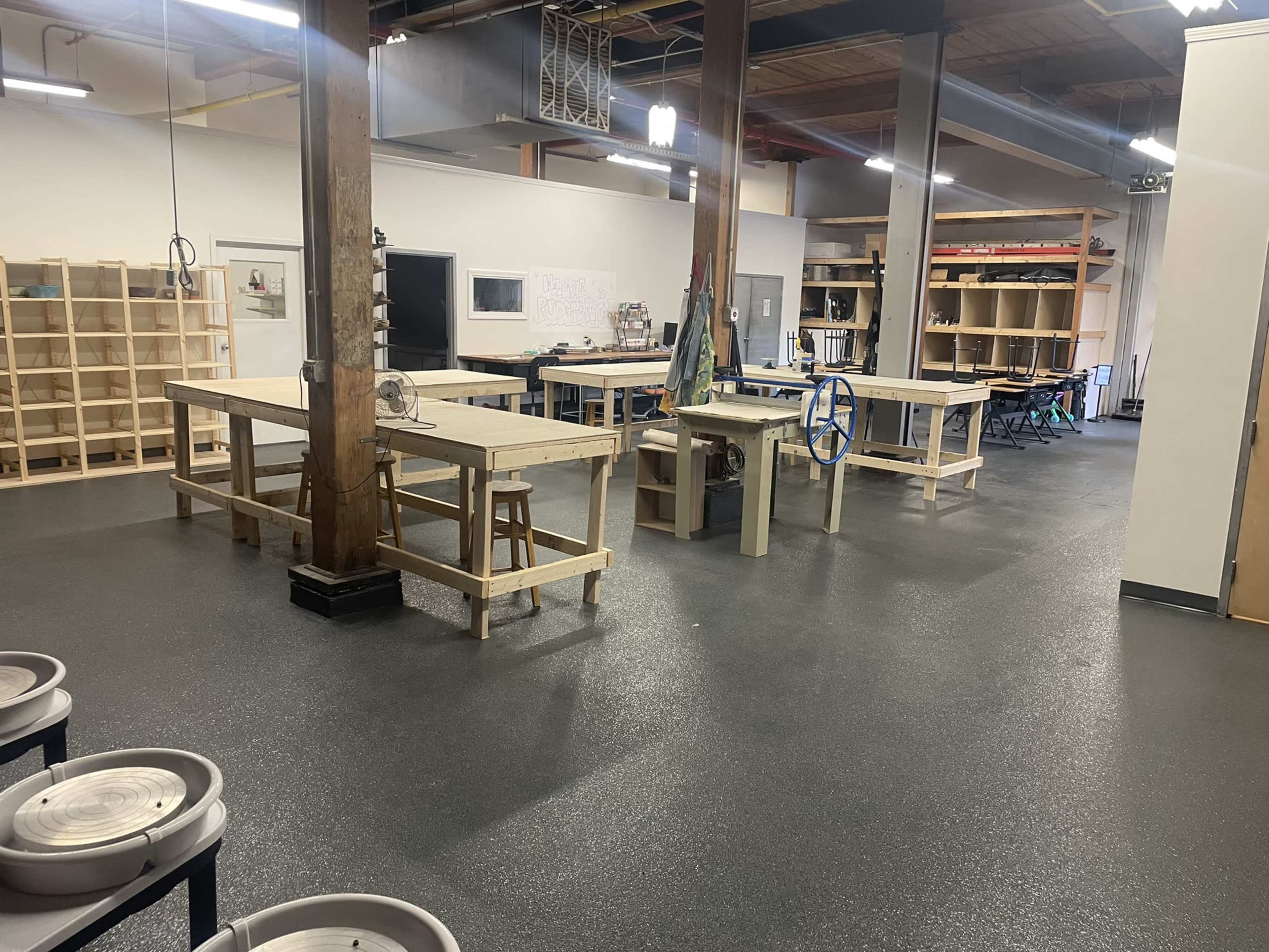An indoor workspace with multiple wooden tables, shelves, and pottery wheels, all arranged neatly in a well-lit room.