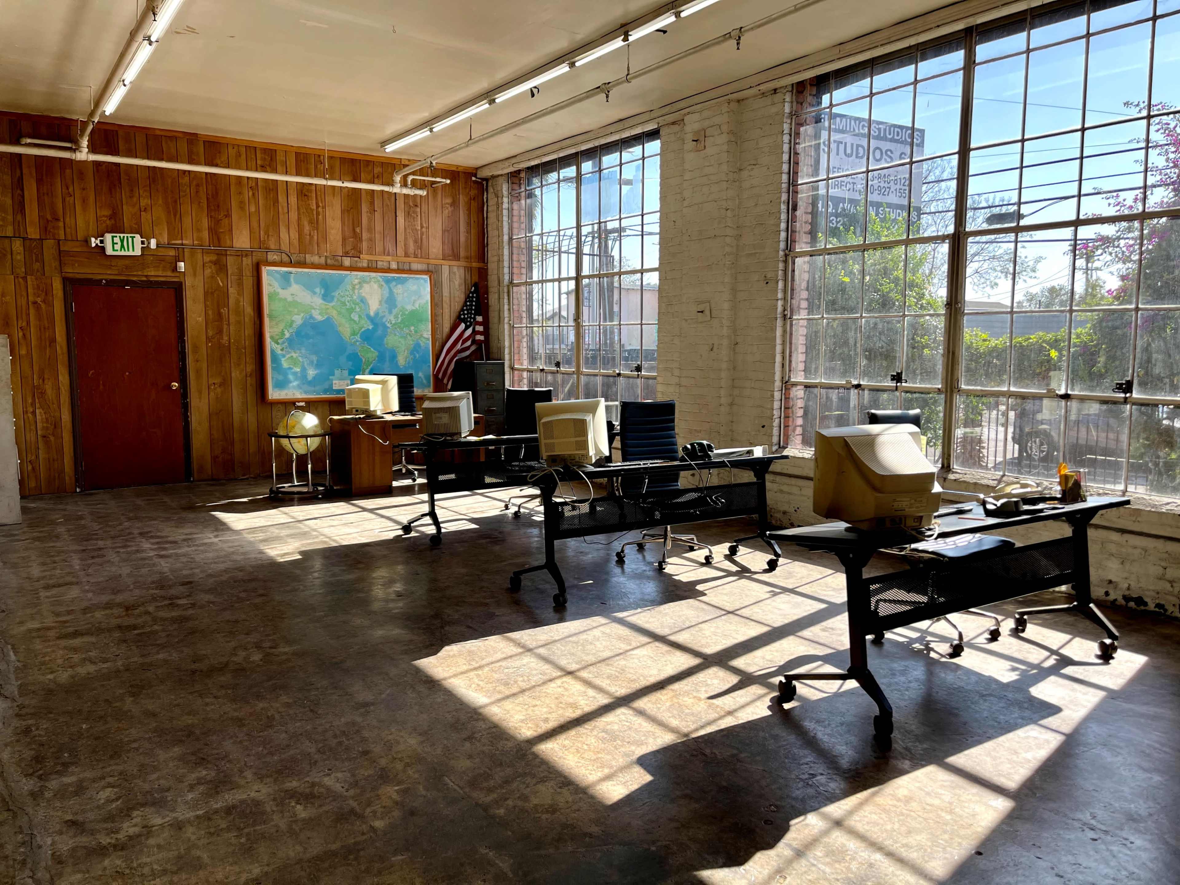 The image depicts an empty office space with multiple desks, old computer monitors, a map on the wall, and large windows allowing natural light to enter.