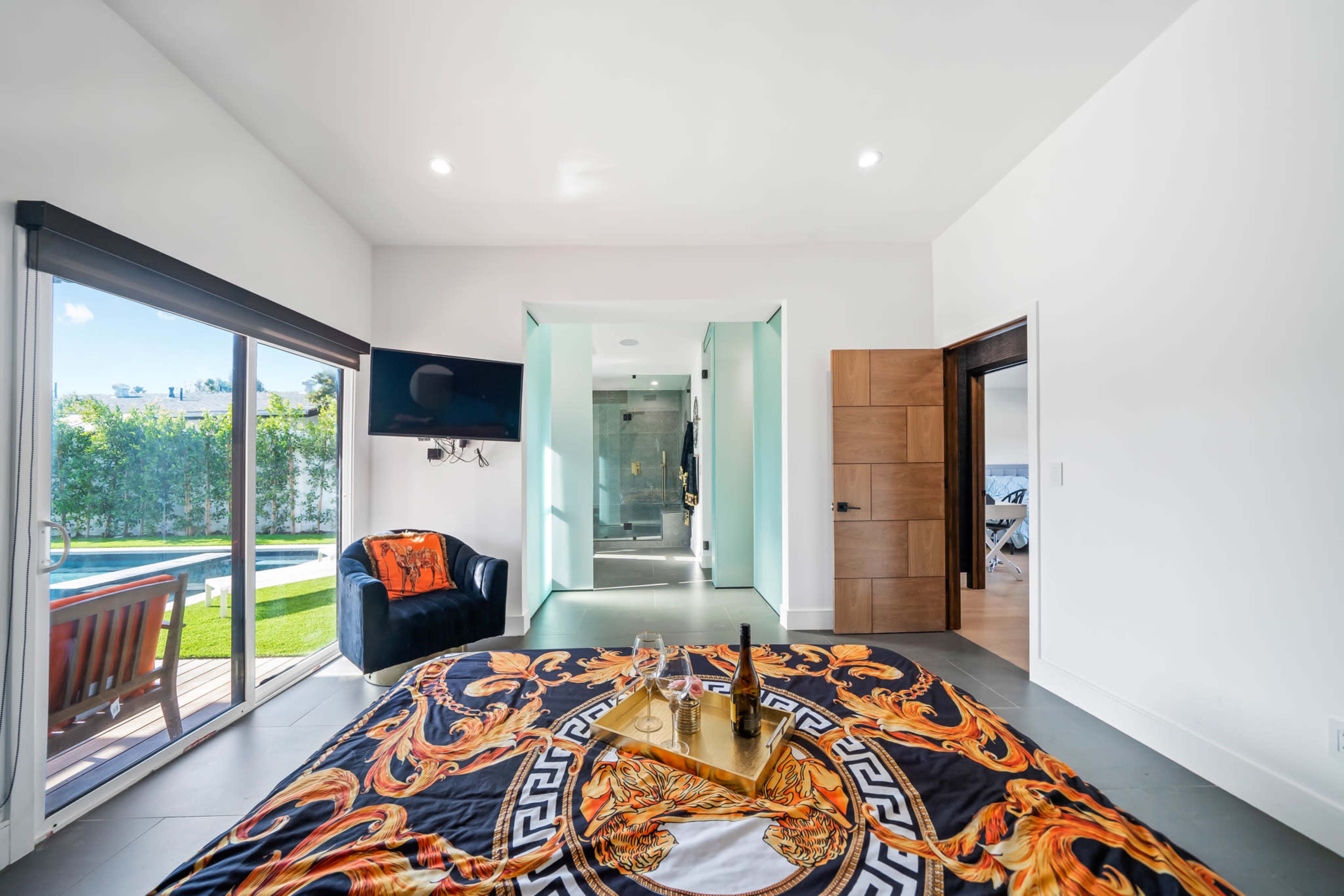 A modern bedroom featuring a large patterned bedspread, a television mounted on the wall, and an ensuite bathroom with glass doors, set against a backdrop of large windows overlooking a pool area.