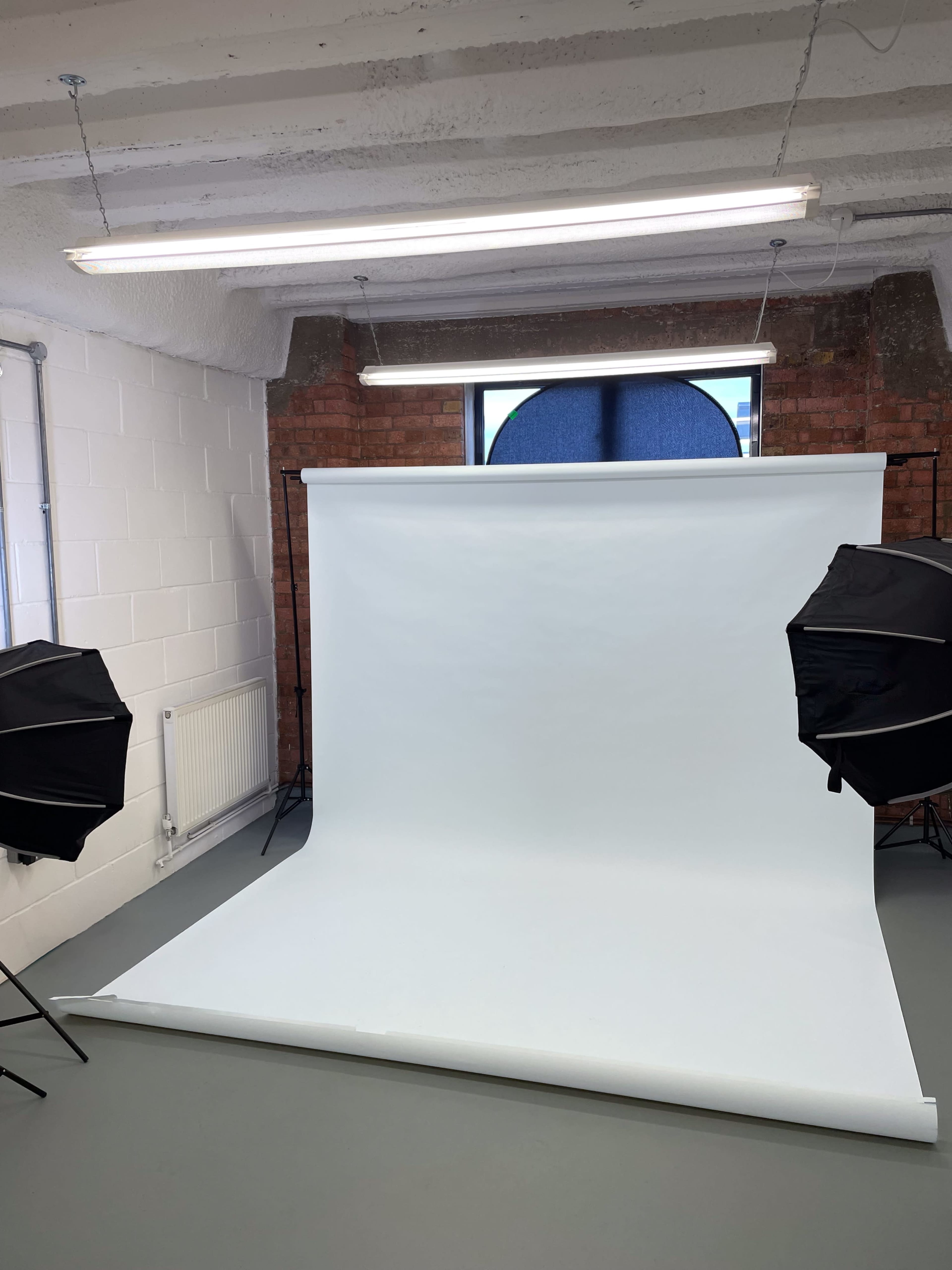 The image shows a photography studio with a white backdrop, two black softbox lights, and a window in the background.